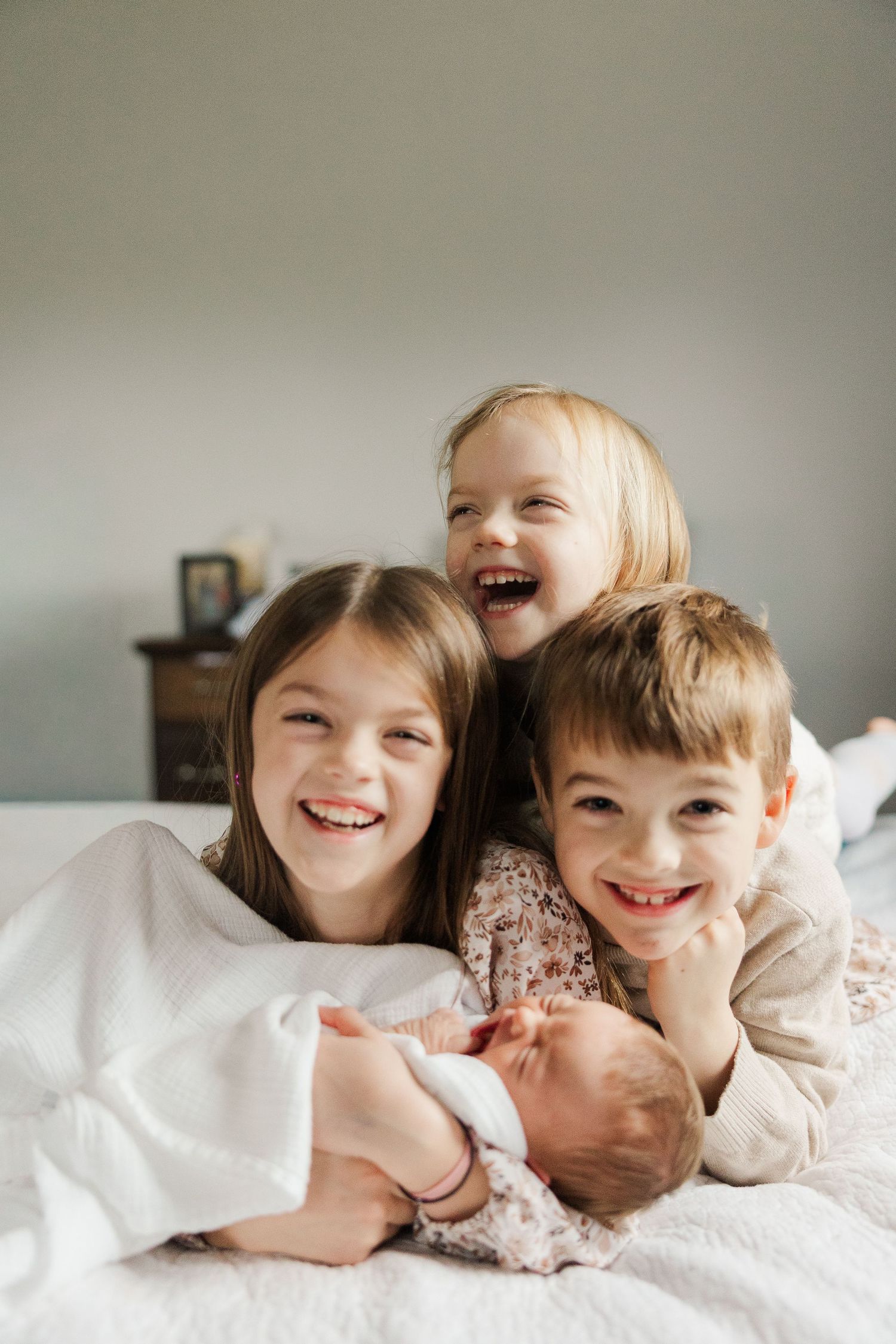 Three smiling children lying on a white bed, with the two older kids cuddling a newborn baby.