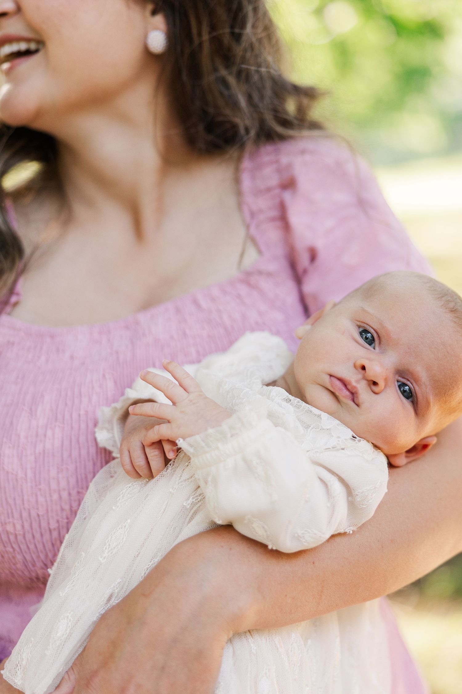 A mother in a pink dress holds her newborn baby dressed in white in a sunlit outdoor setting.