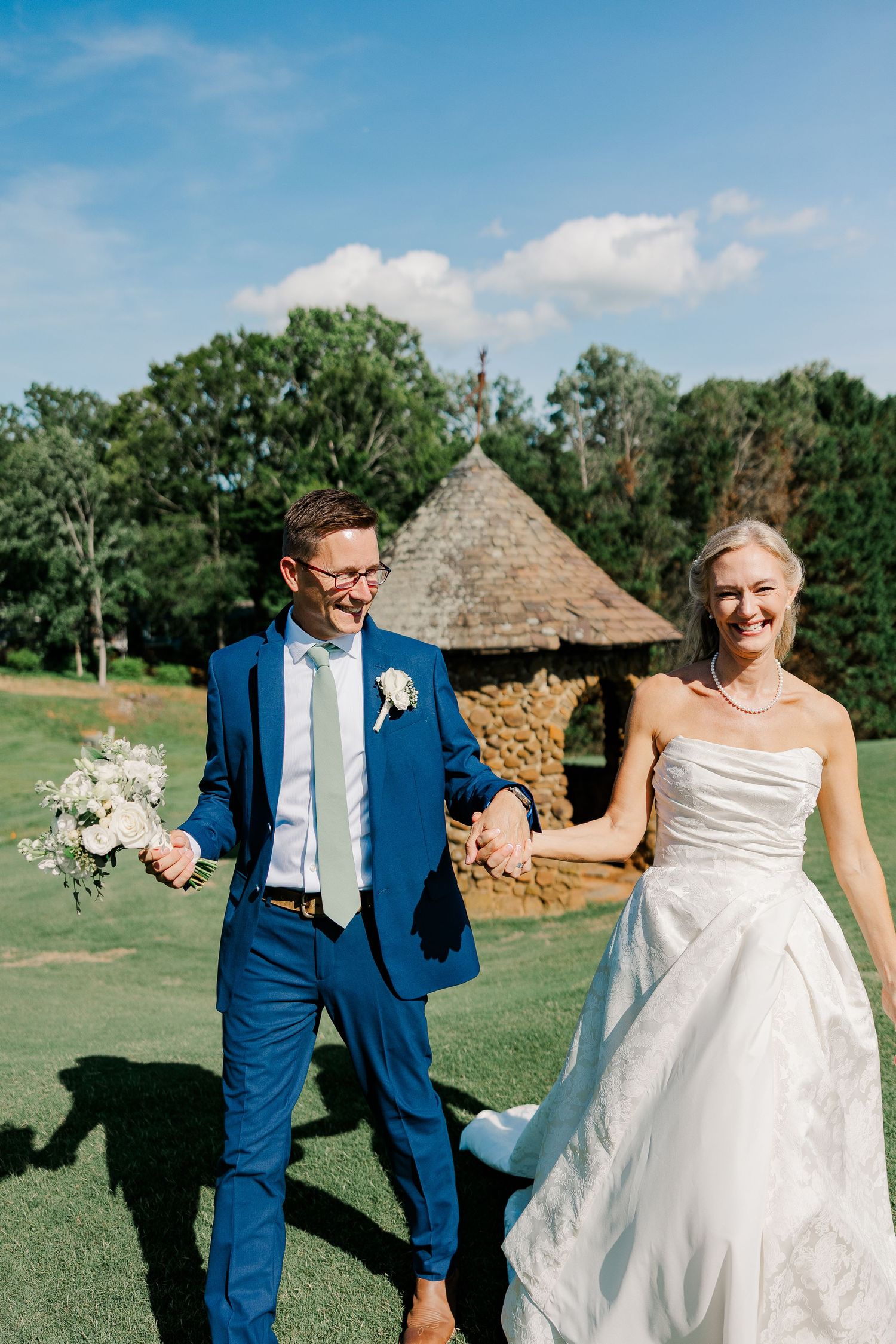 Smiling bride in a strapless gown and groom in a blue suit holding hands and a bouquet near a stone gazebo on a sunny wedding day.