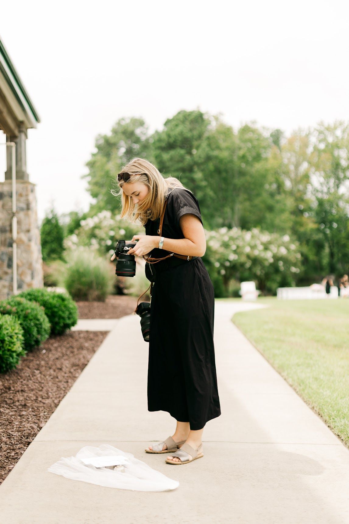 Woman in black dress reviewing photos on camera while standing on a garden pathway outdoors.