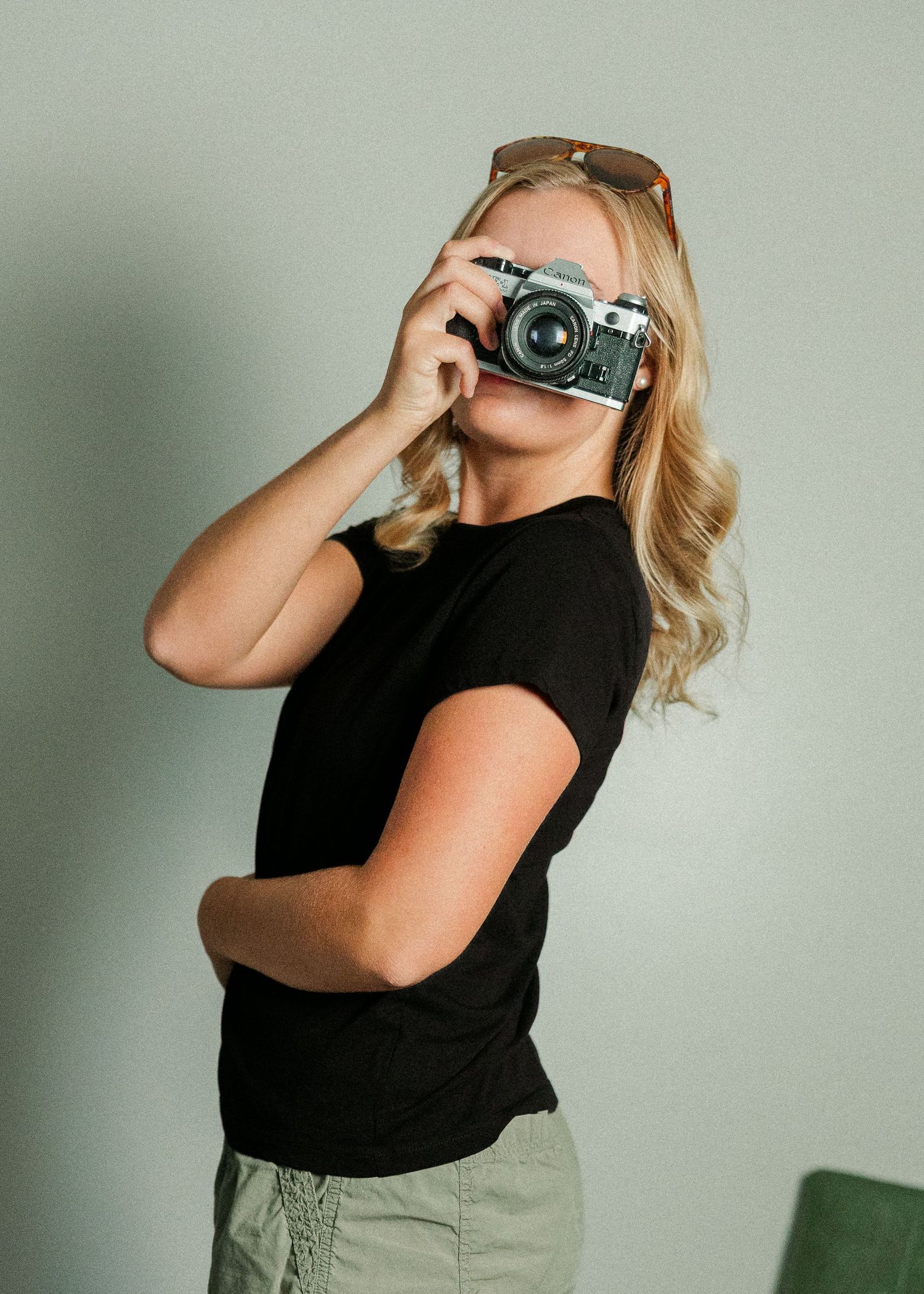 Blonde woman in black t-shirt holding a vintage film camera up to her face with sunglasses on her head.