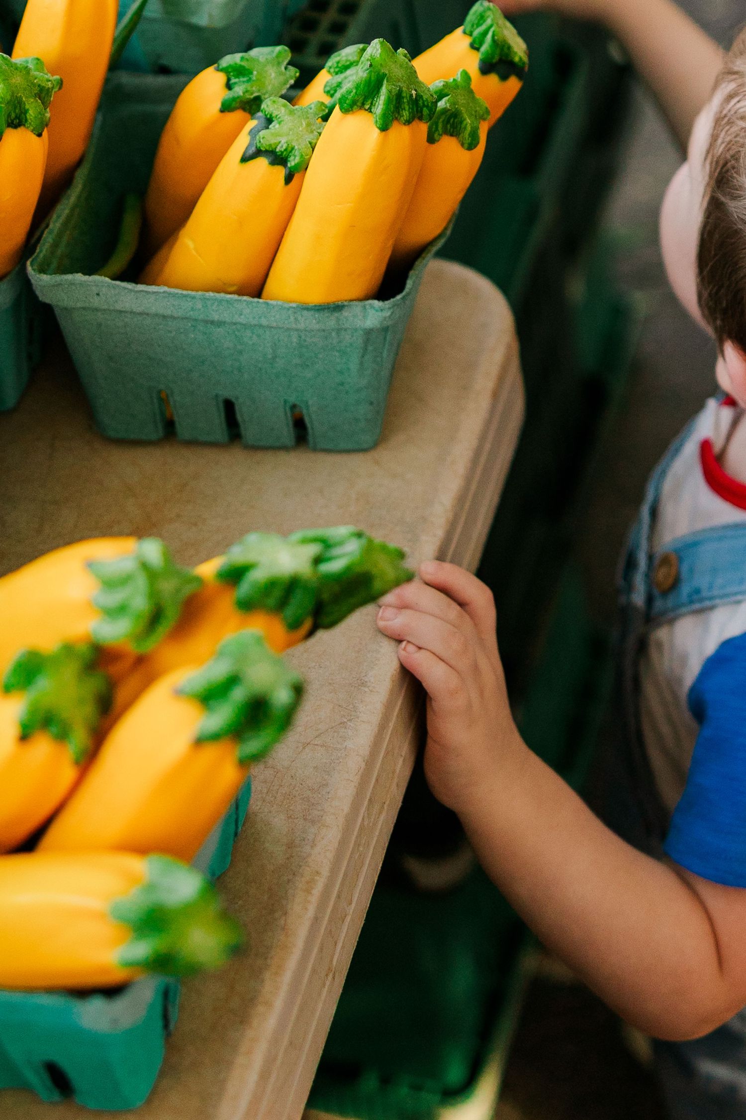 Young child reaching for bright yellow zucchini squash displayed in green berry baskets at a farmers market stand.