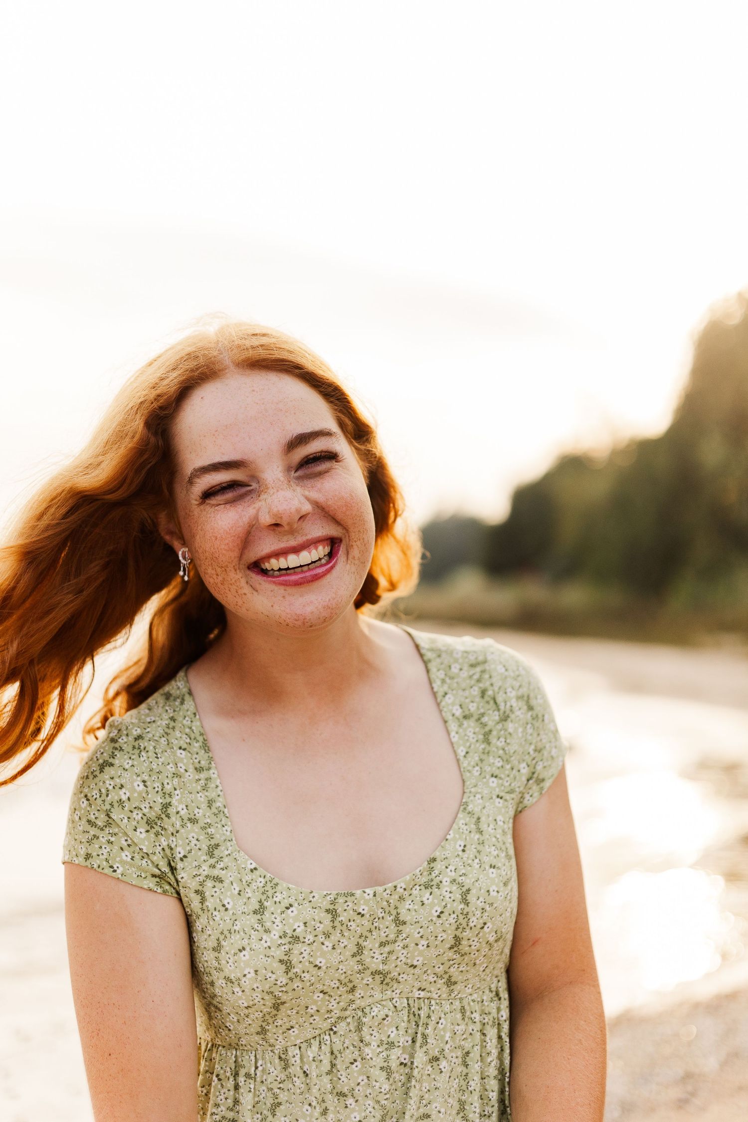 Smiling young woman with red hair and freckles wearing a green floral dress outdoors in golden sunlight.