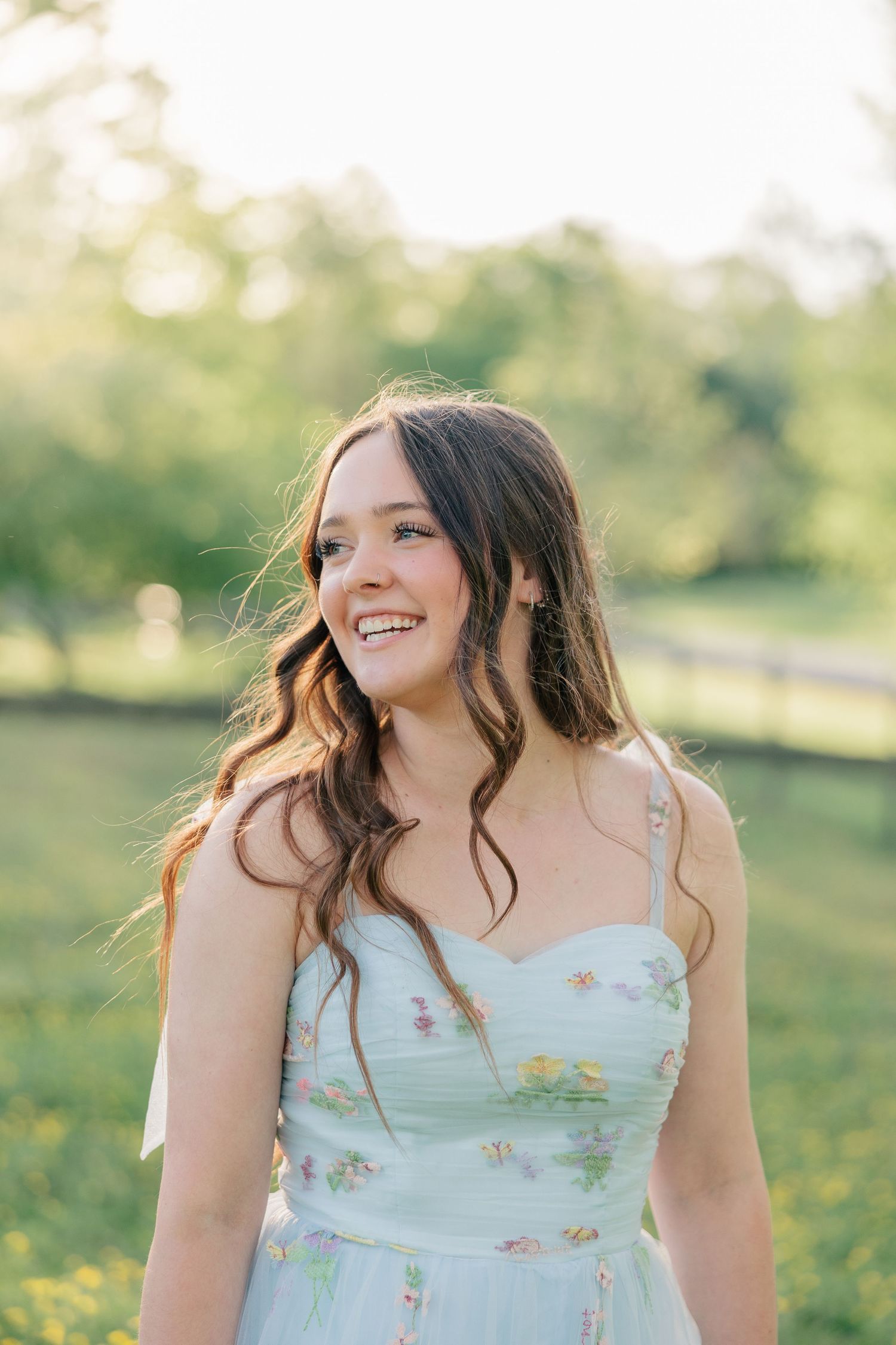 Smiling young woman in a floral mint dress standing in a sunlit green field with trees in the background.
