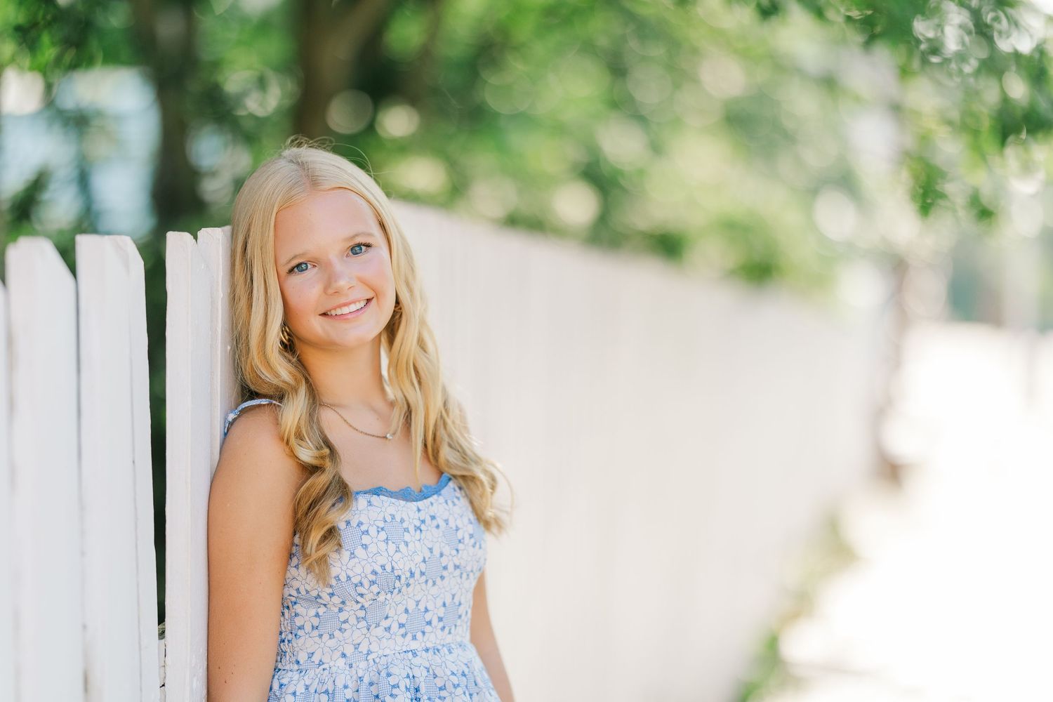 Smiling blonde girl in blue floral dress leaning against white picket fence outdoors.