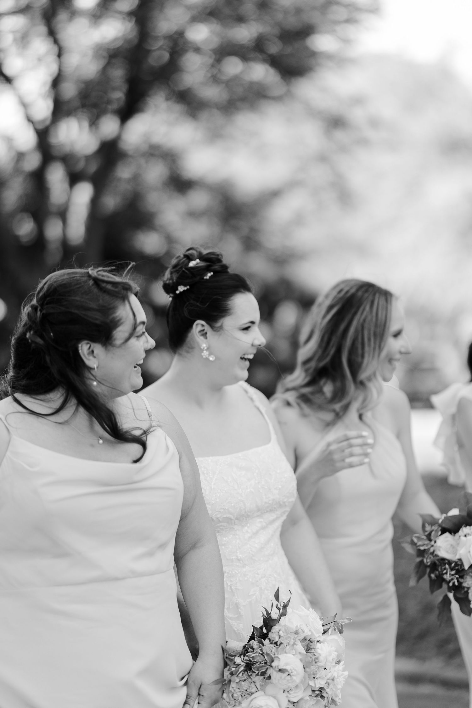 Black and white photo of three laughing bridesmaids in elegant dresses walking outdoors, one holding a white floral bouquet.