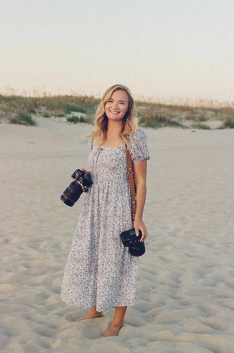 A smiling blonde woman in a floral midi dress holds two cameras while standing barefoot on a sandy beach at sunset.