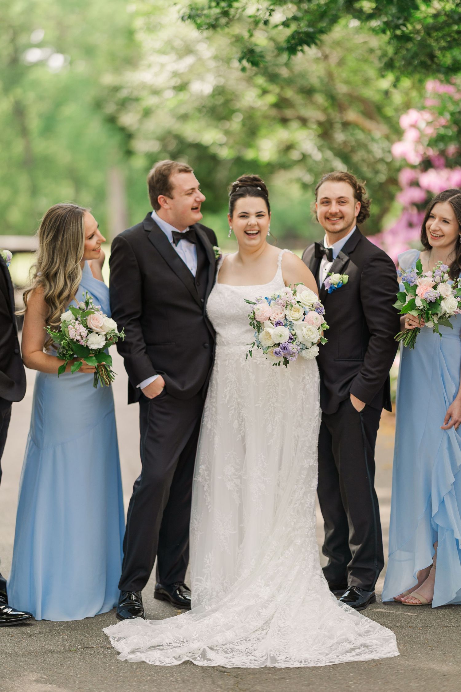 Smiling bride in a lace gown holding a floral bouquet poses outdoors with two groomsmen in dark suits and two bridesmaids in blue dresses.