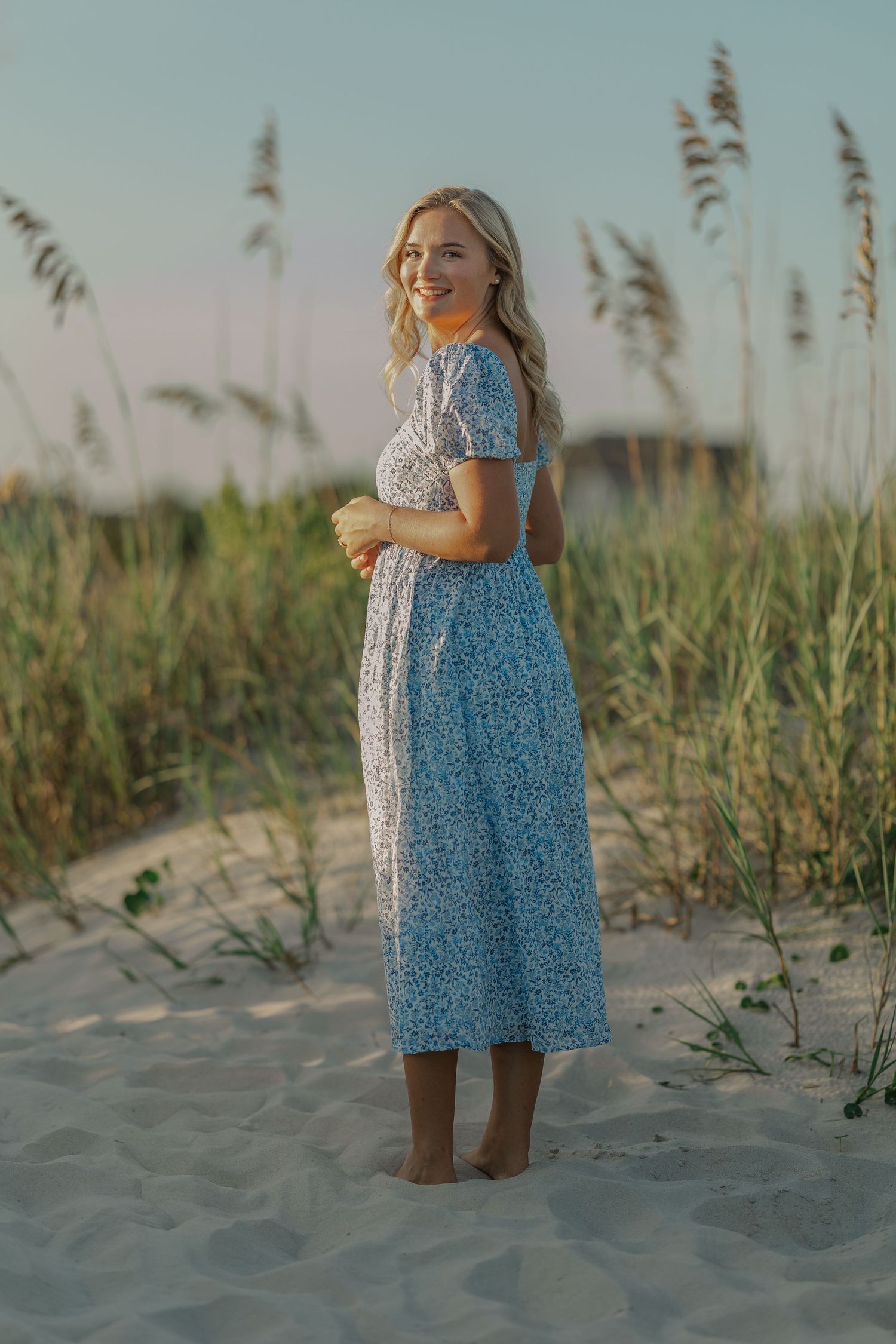 Young woman in blue floral midi dress standing barefoot on sandy beach dunes with sea oats at sunset.