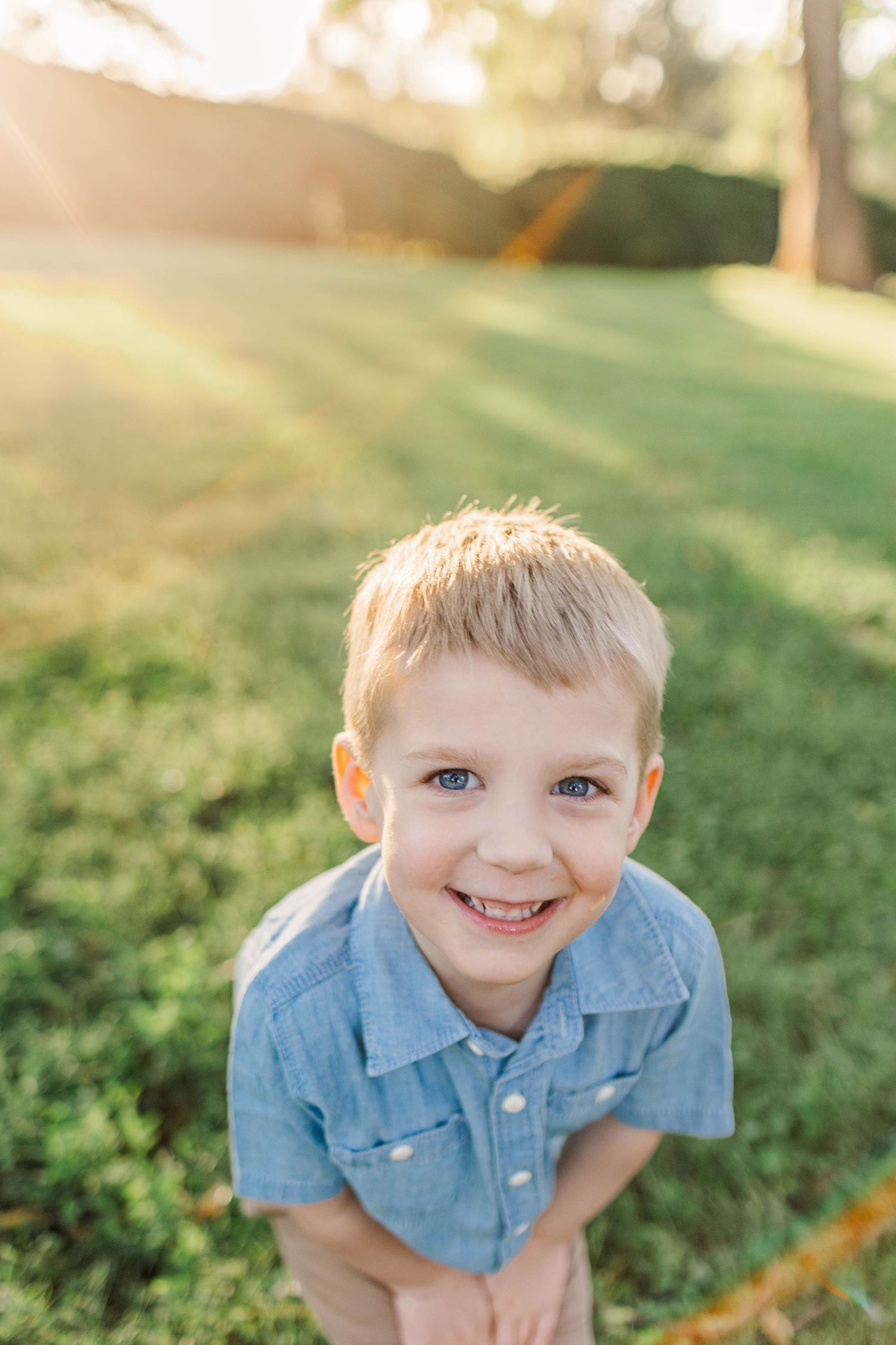 Smiling blond boy with blue eyes in a blue shirt, leaning forward on a sunny green lawn.