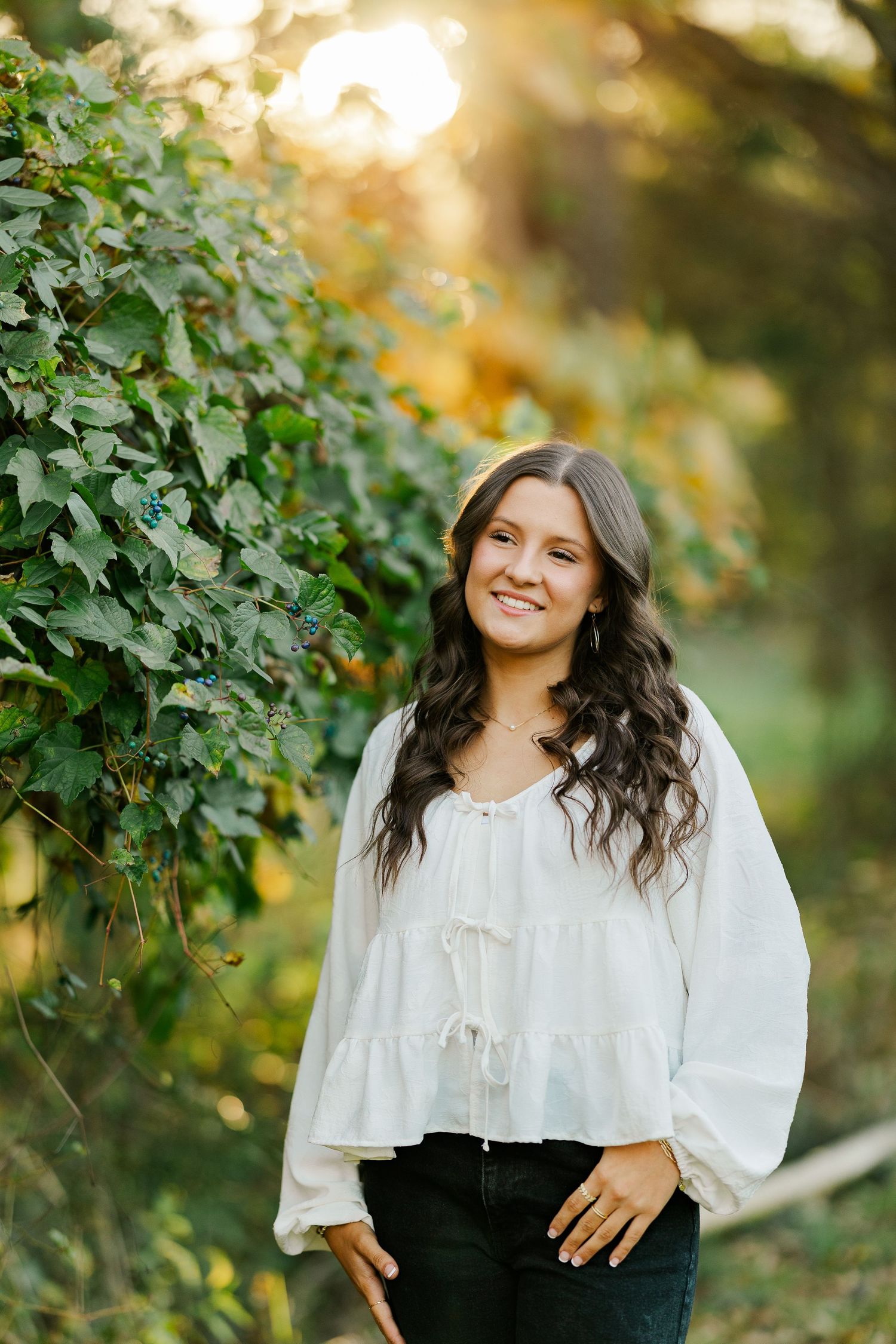 Young woman with long wavy hair in white blouse and jeans smiling outdoors among lush green vines at golden hour.
