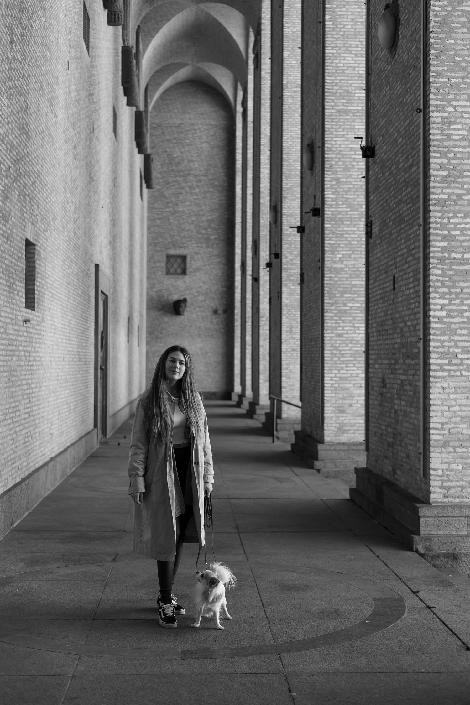 A person stands with a small white dog in an arched corridor with brick walls and dramatic shadows.