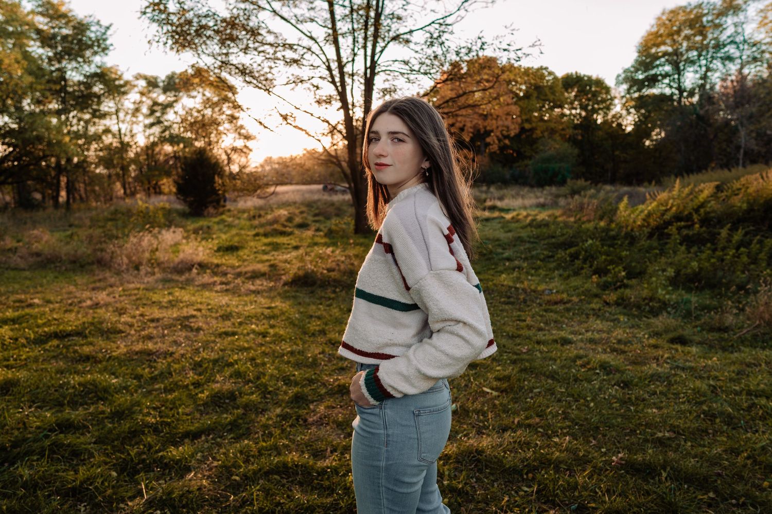 Young woman in cream sweater and jeans poses in golden hour sunlight in a lush green field with autumn trees.