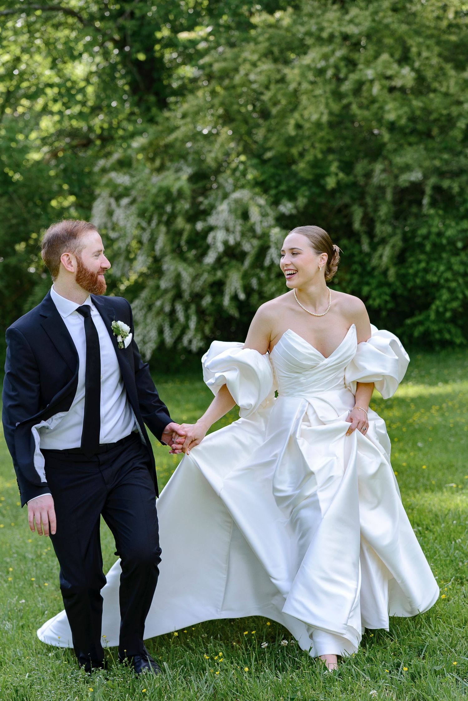 A joyful bride in a white off-shoulder gown and groom in black suit hold hands walking through a lush green garden.