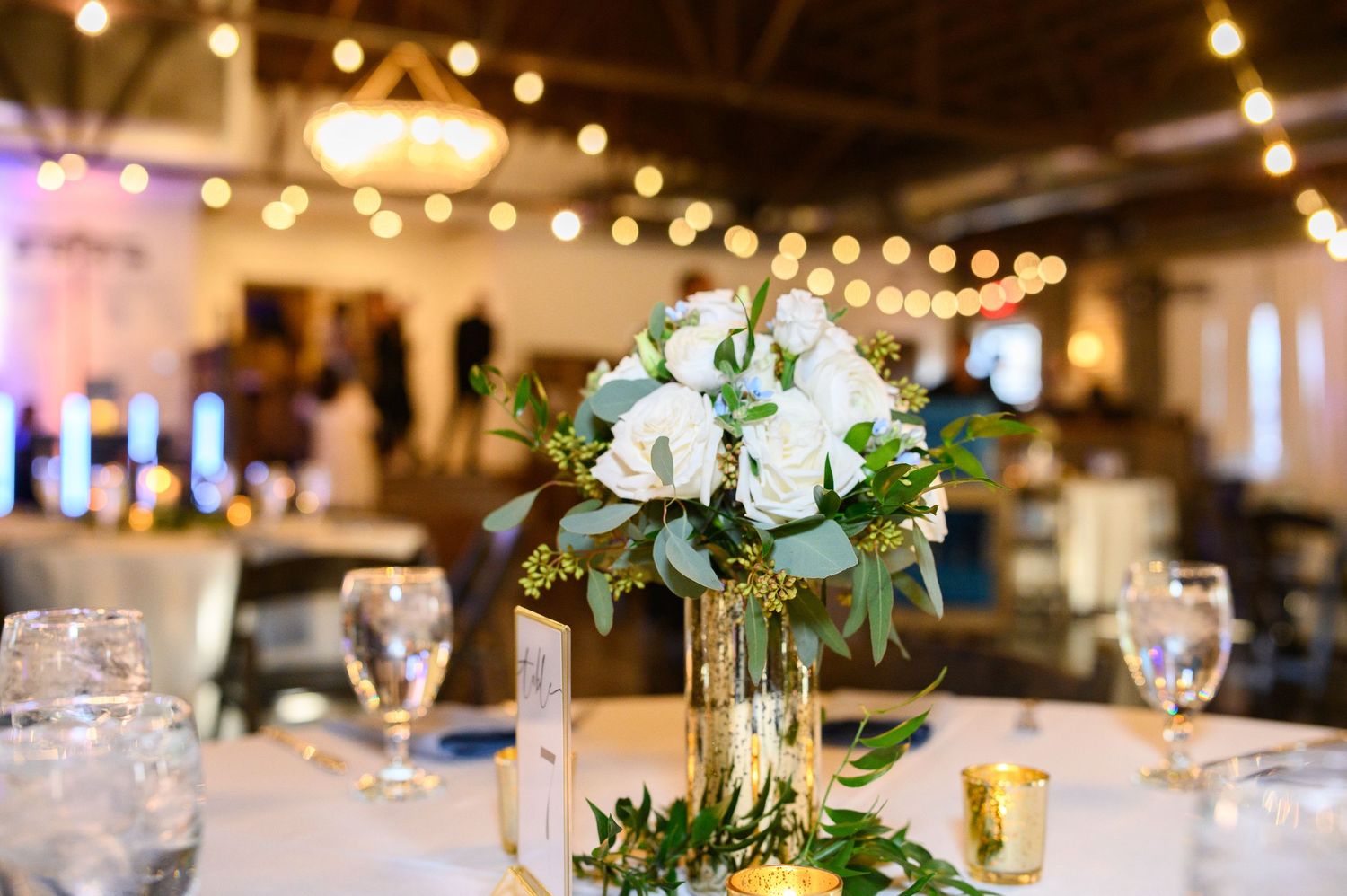 Elegant wedding centerpiece with white flowers and greenery on table with string lights and glassware in reception venue.
