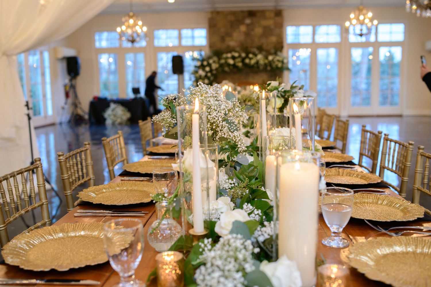 Elegant wedding reception table setting with gold chargers, white candles and floral garland runner in bright venue setting.