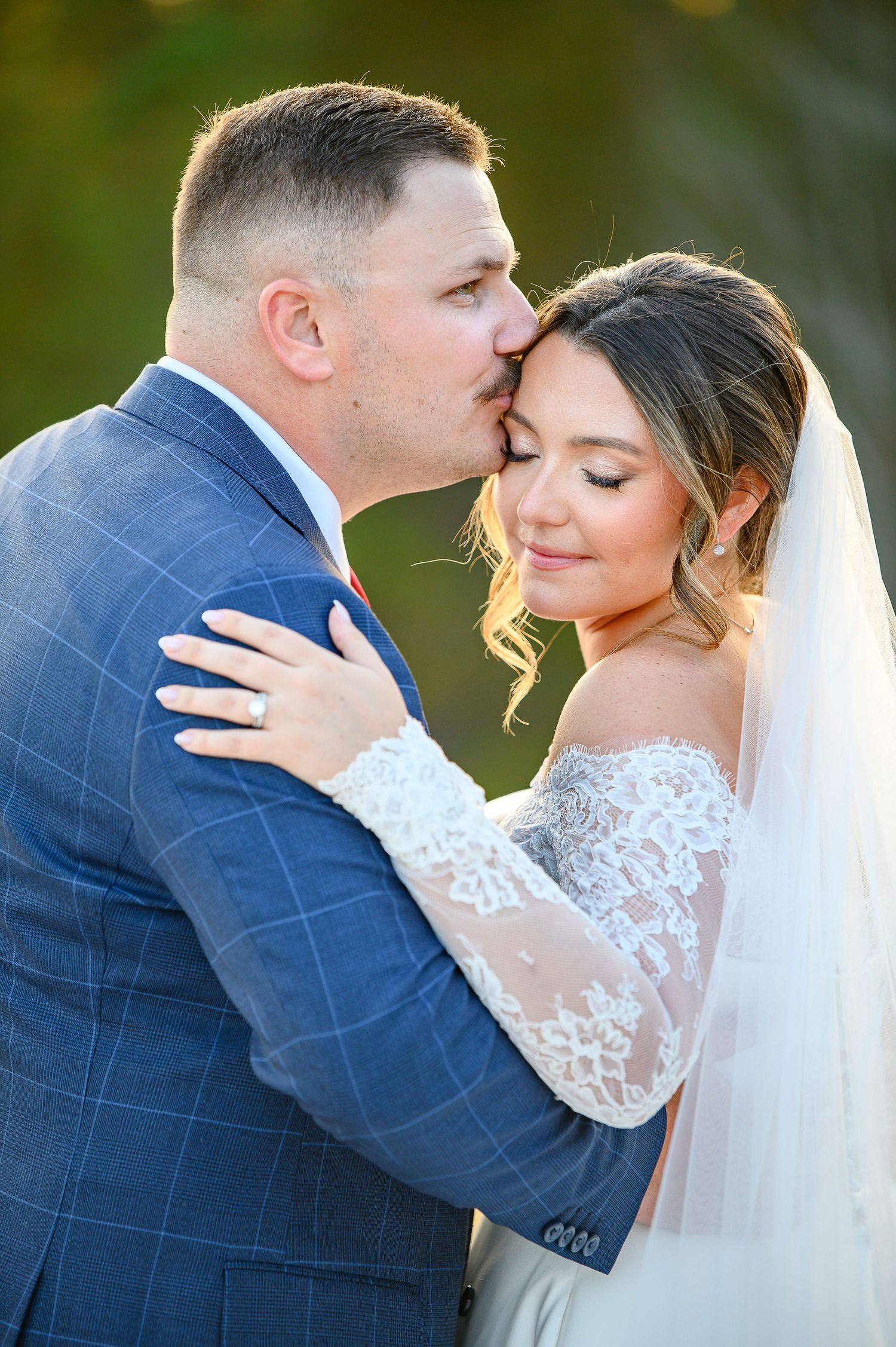 Groom in blue suit kisses bride on forehead as she smiles in lace off-shoulder wedding gown and veil.