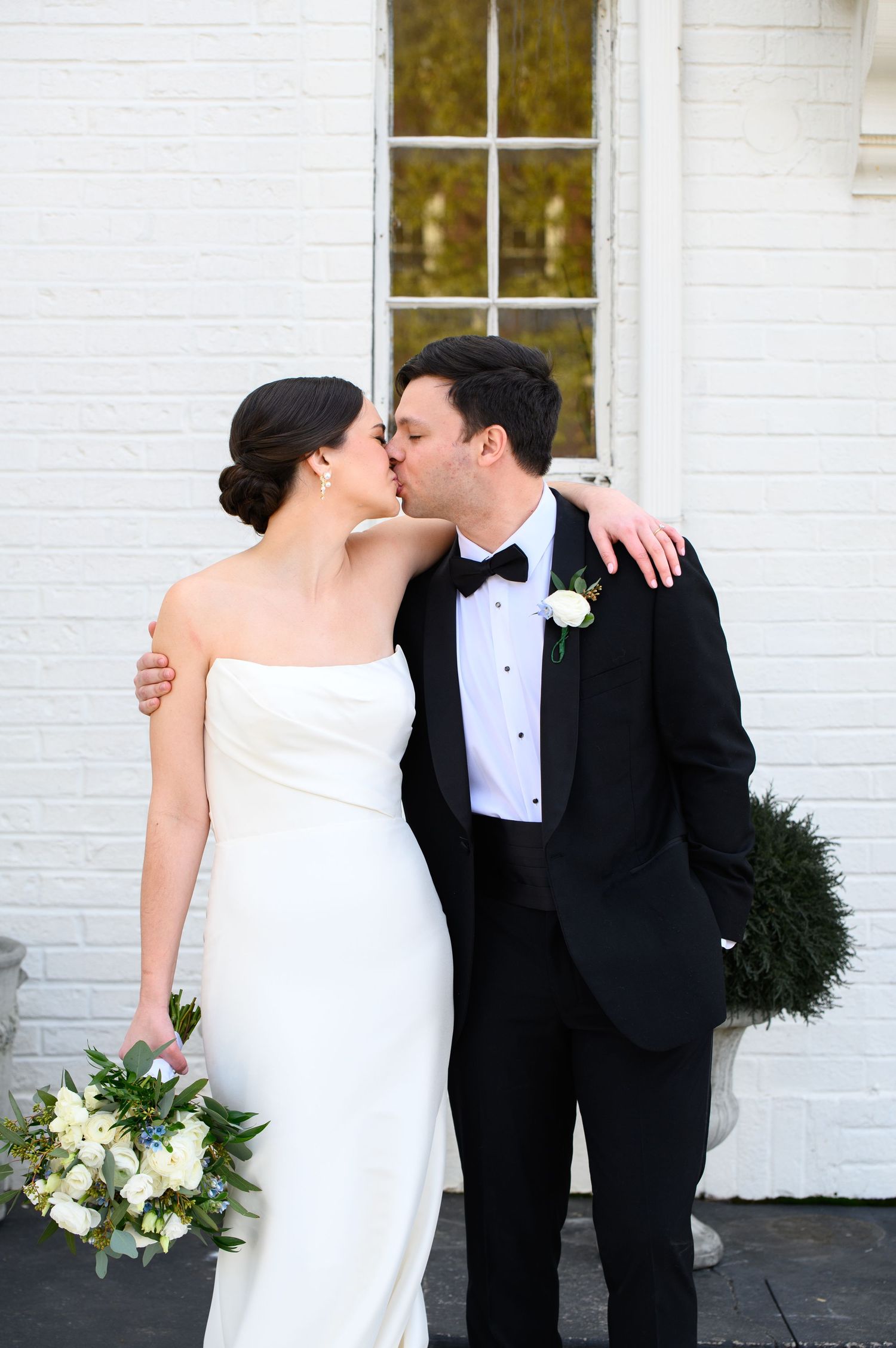 An elegant couple in formal wedding attire share a kiss outside a white brick building with a floral bouquet.