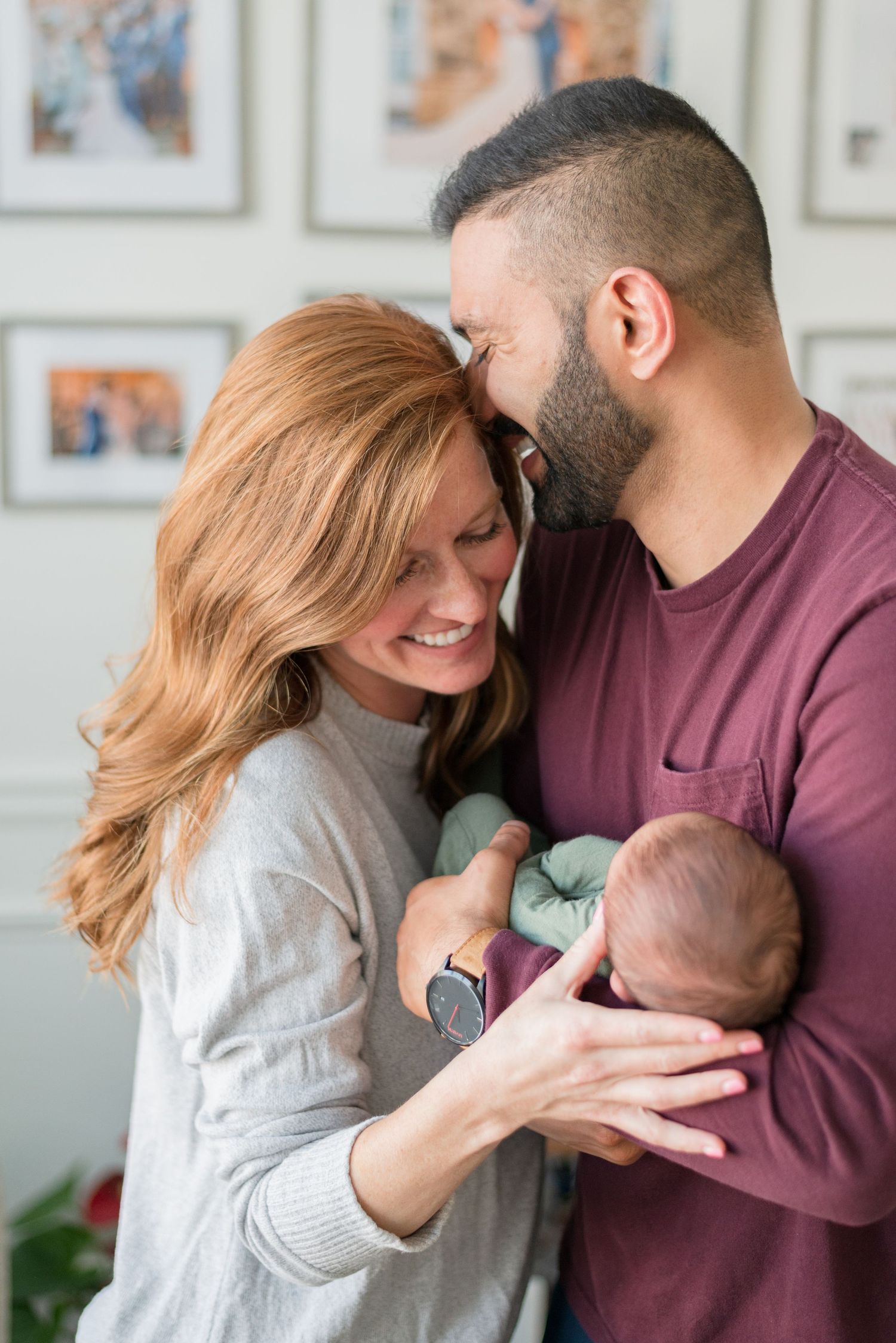 Parents share intimate moment while holding their newborn baby against wall with framed photos in background.