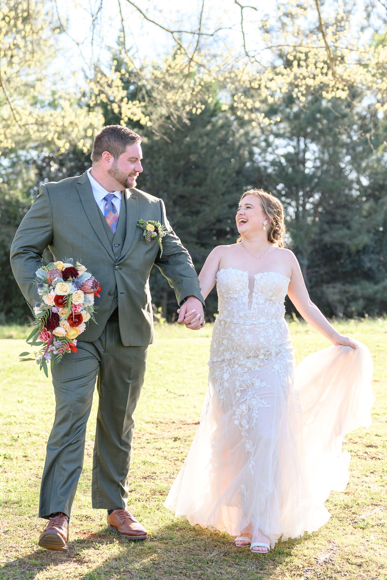 Happy bride and groom walk hand-in-hand outdoors, she in a floral gown holding her skirt, he in a gray suit with bouquet.