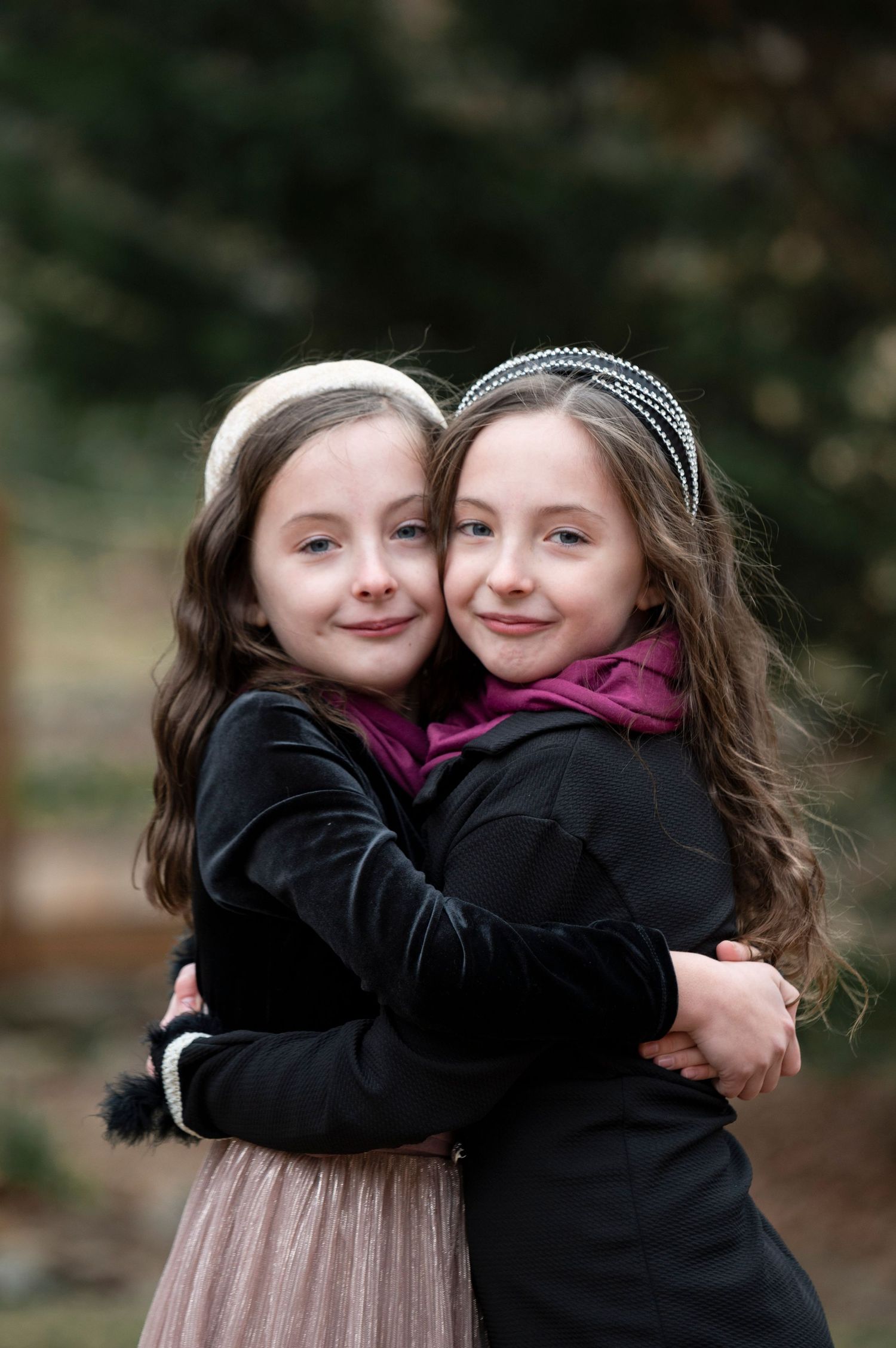 Two young girls in black coats share a warm embrace while smiling at the camera outdoors. family photos in canton, ga
