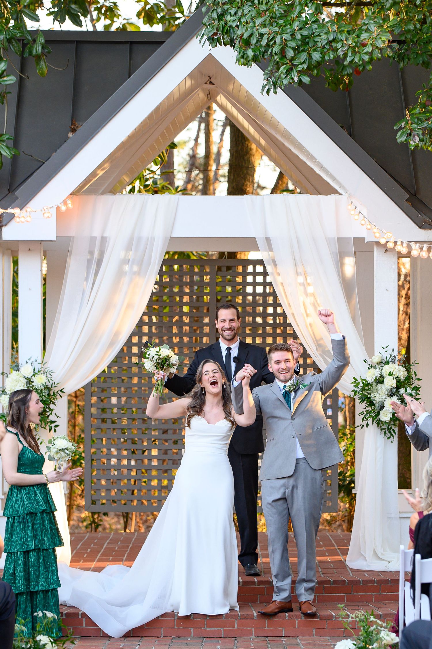 Joyful celebration under a white draped wedding arch decorated with string lights and floral arrangements at an outdoor ceremony.