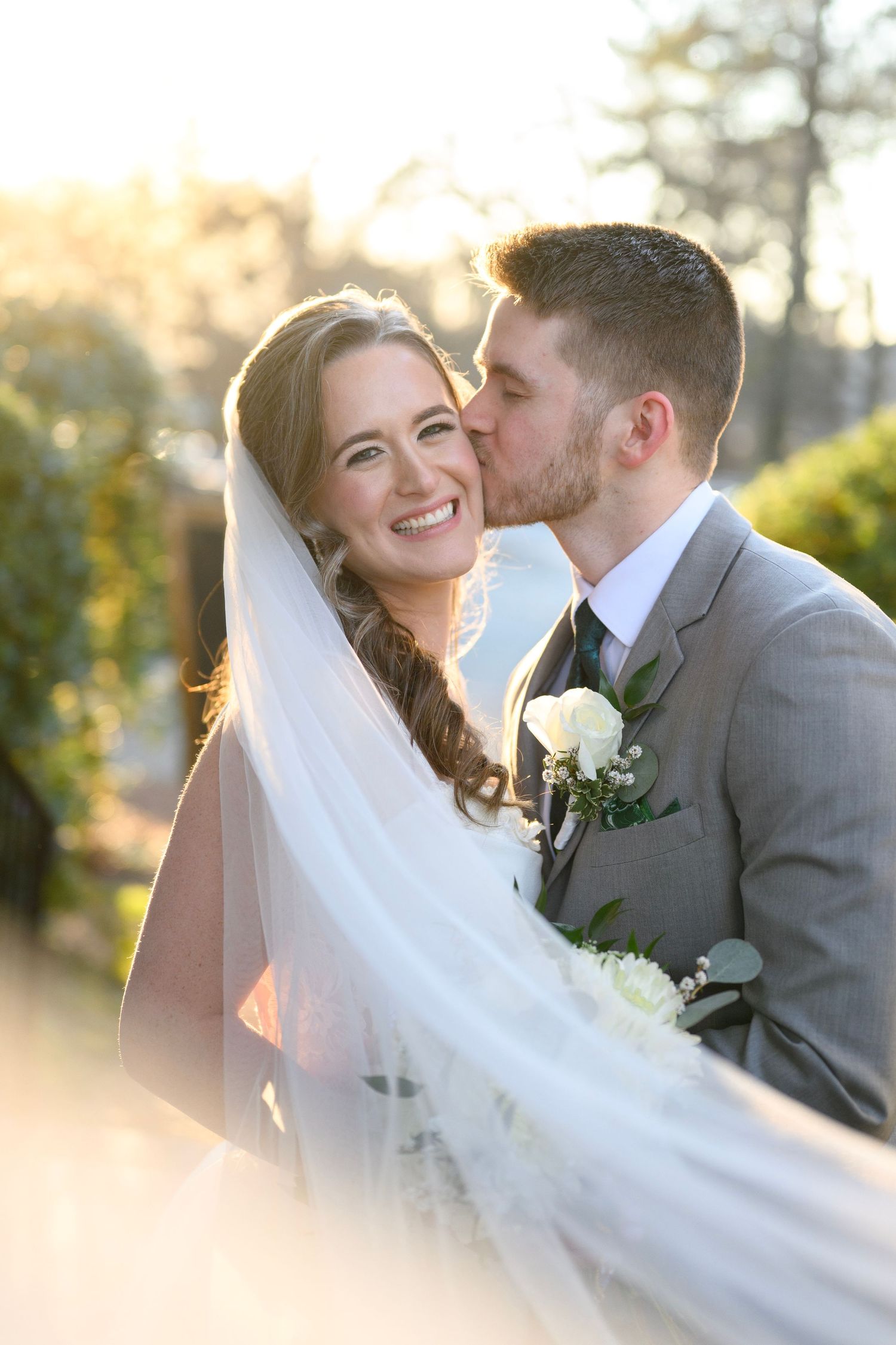 A romantic wedding moment captured in sunlight as a couple embraces with the bride's veil flowing in the breeze.