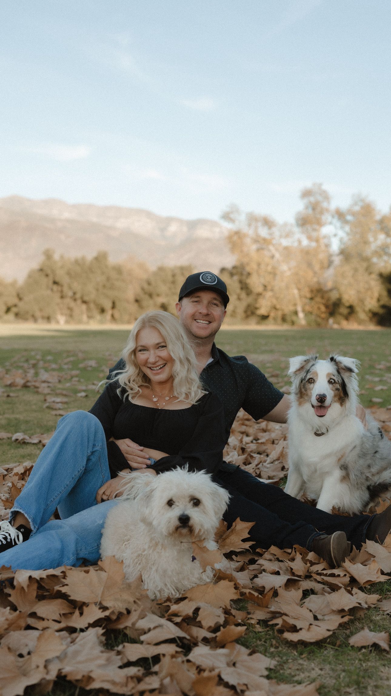 A couple sits in autumn leaves with their three fluffy white dogs enjoying a fall photoshoot outdoors.