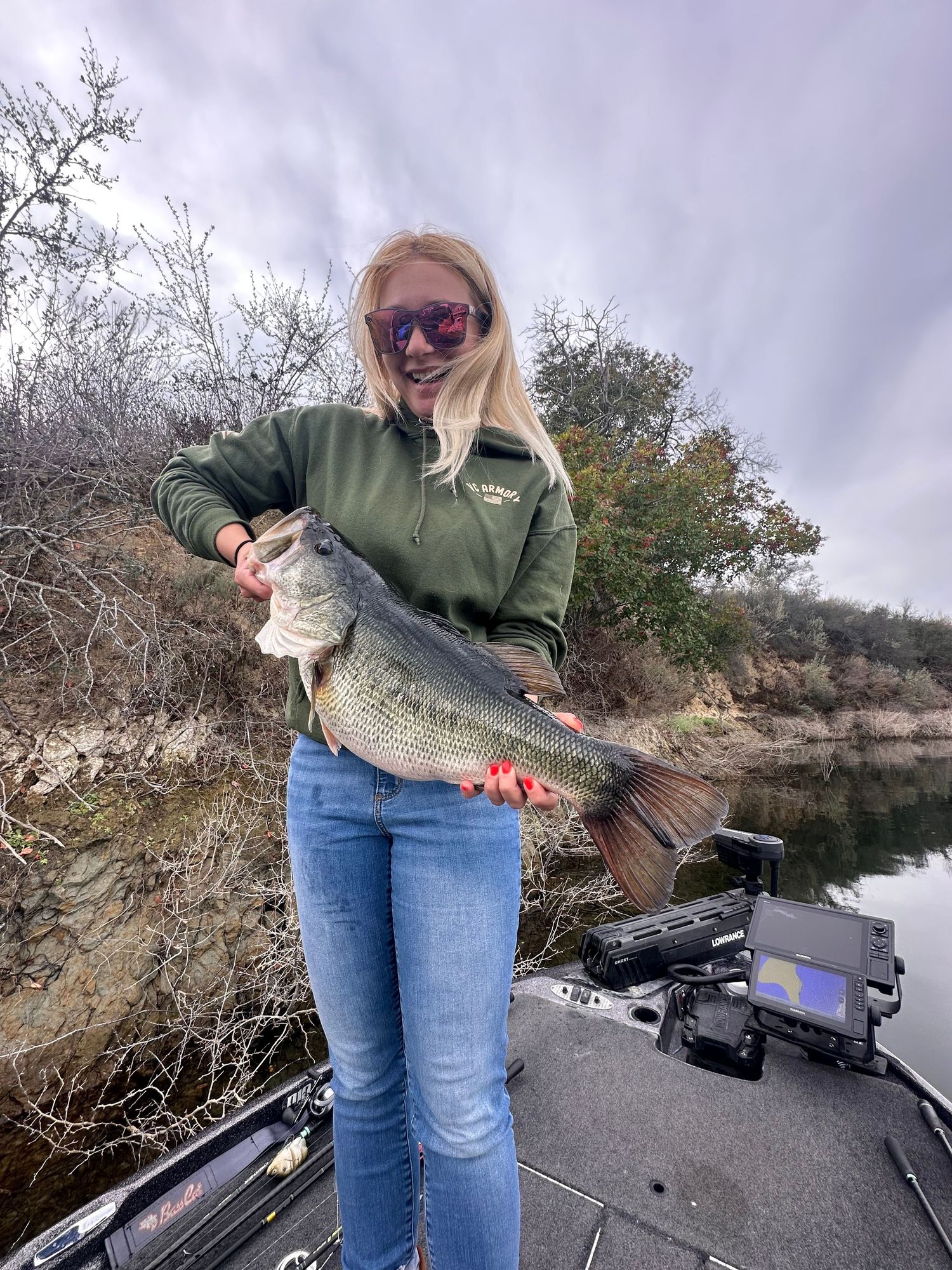 Angler in green sweatshirt and jeans holds up impressive largemouth bass catch while standing on fishing boat at lake.