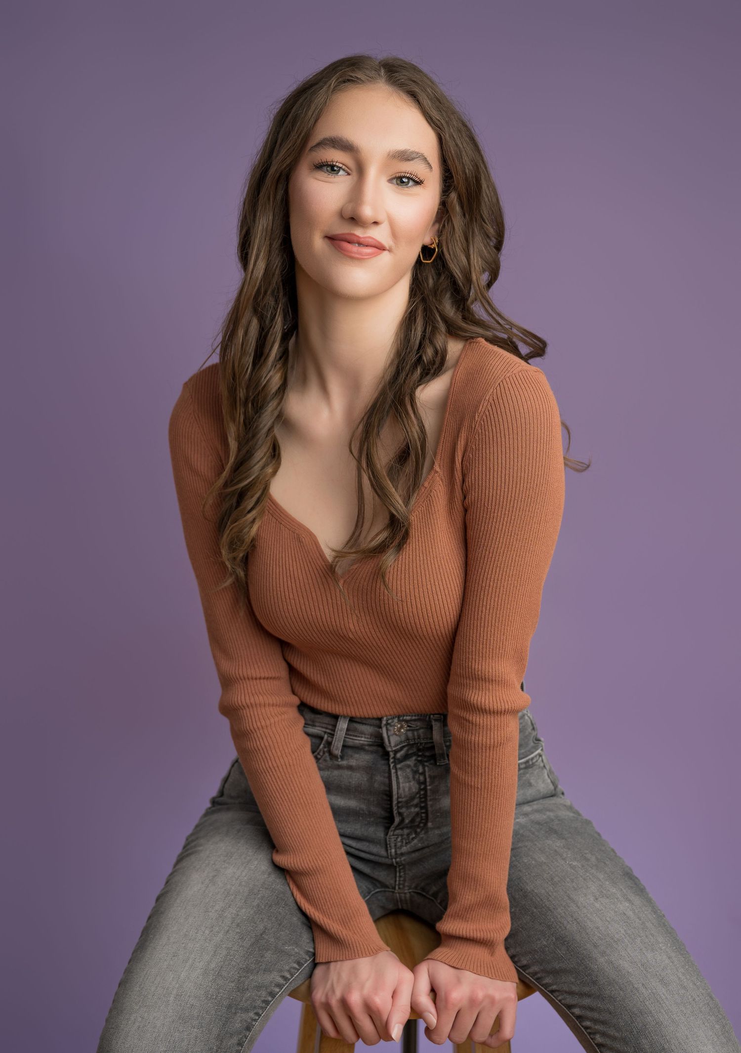 A young person in a brown ribbed sweater and jeans sits against a purple studio background, with long wavy hair framing their face.
