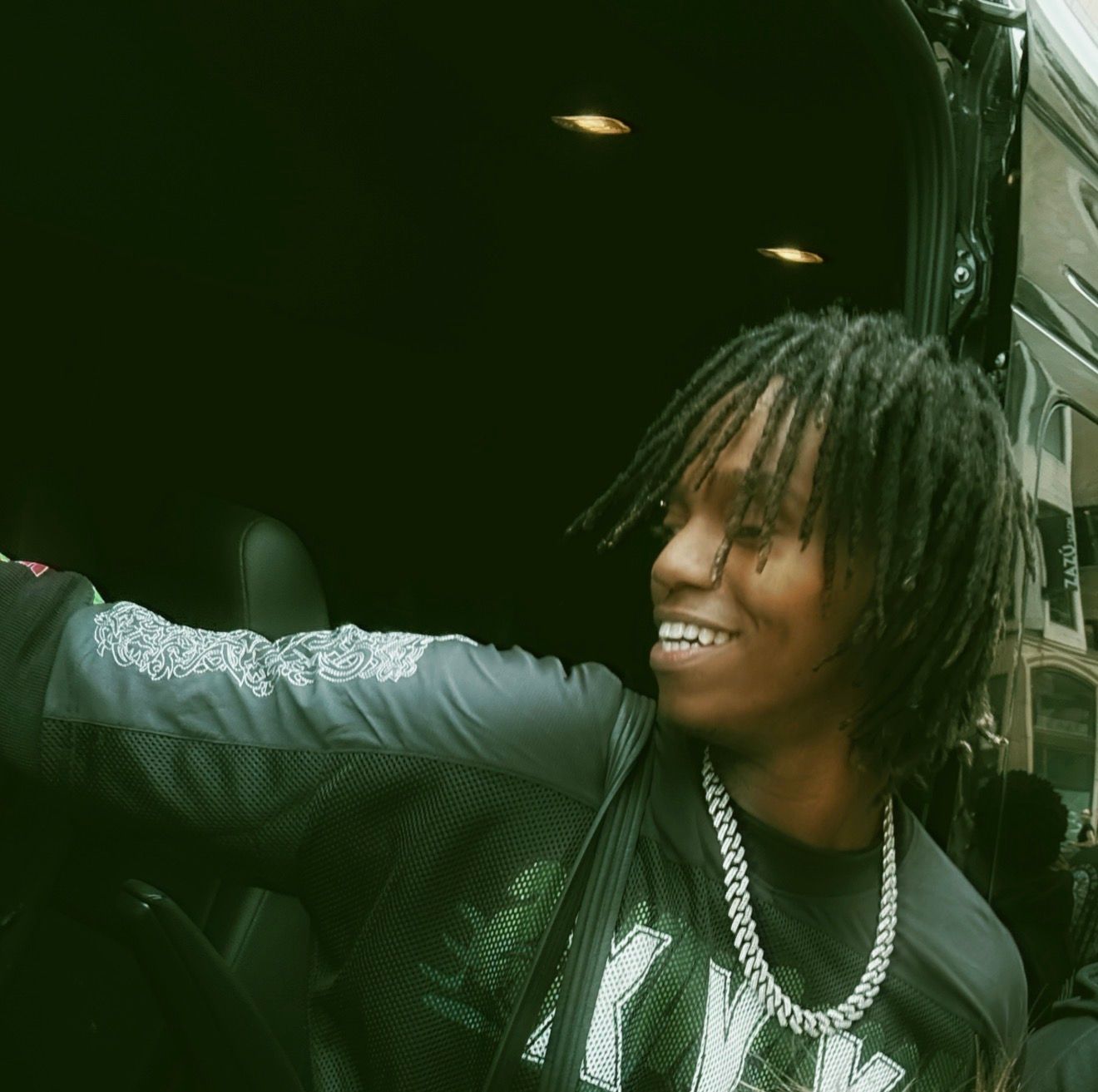 Young man with dreadlocks wearing a chain necklace and black jacket smiling outdoors.