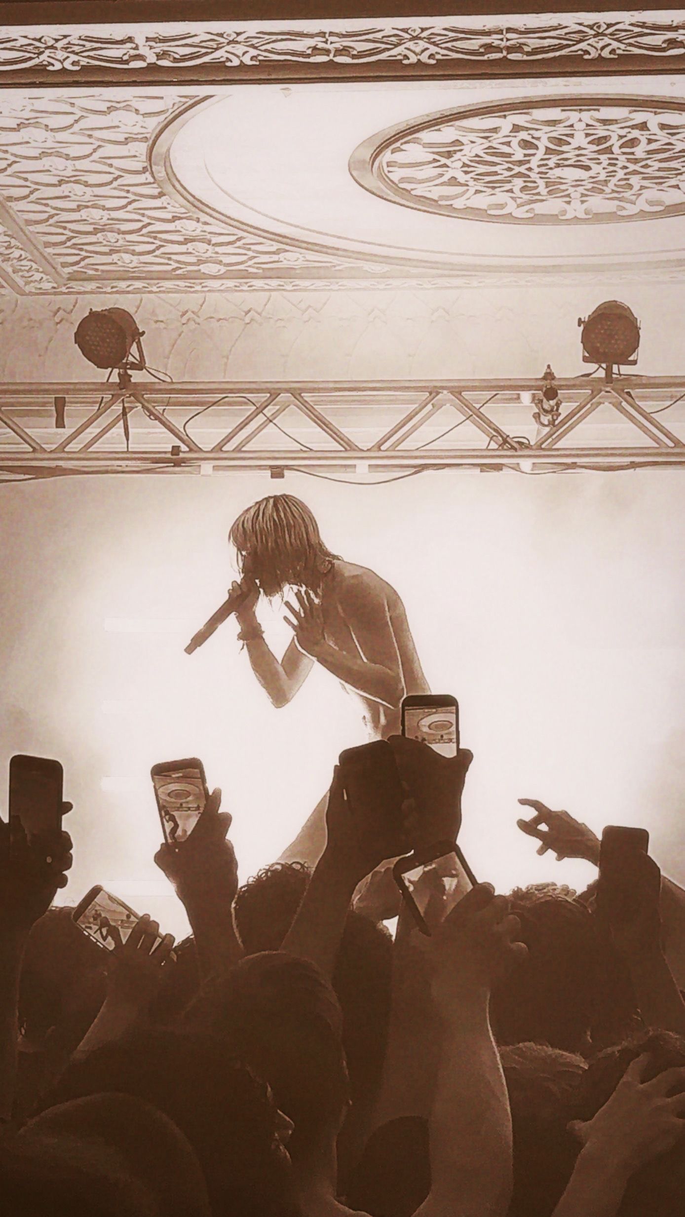 Concert performer singing into microphone above crowd with phones raised in sepia-toned venue.