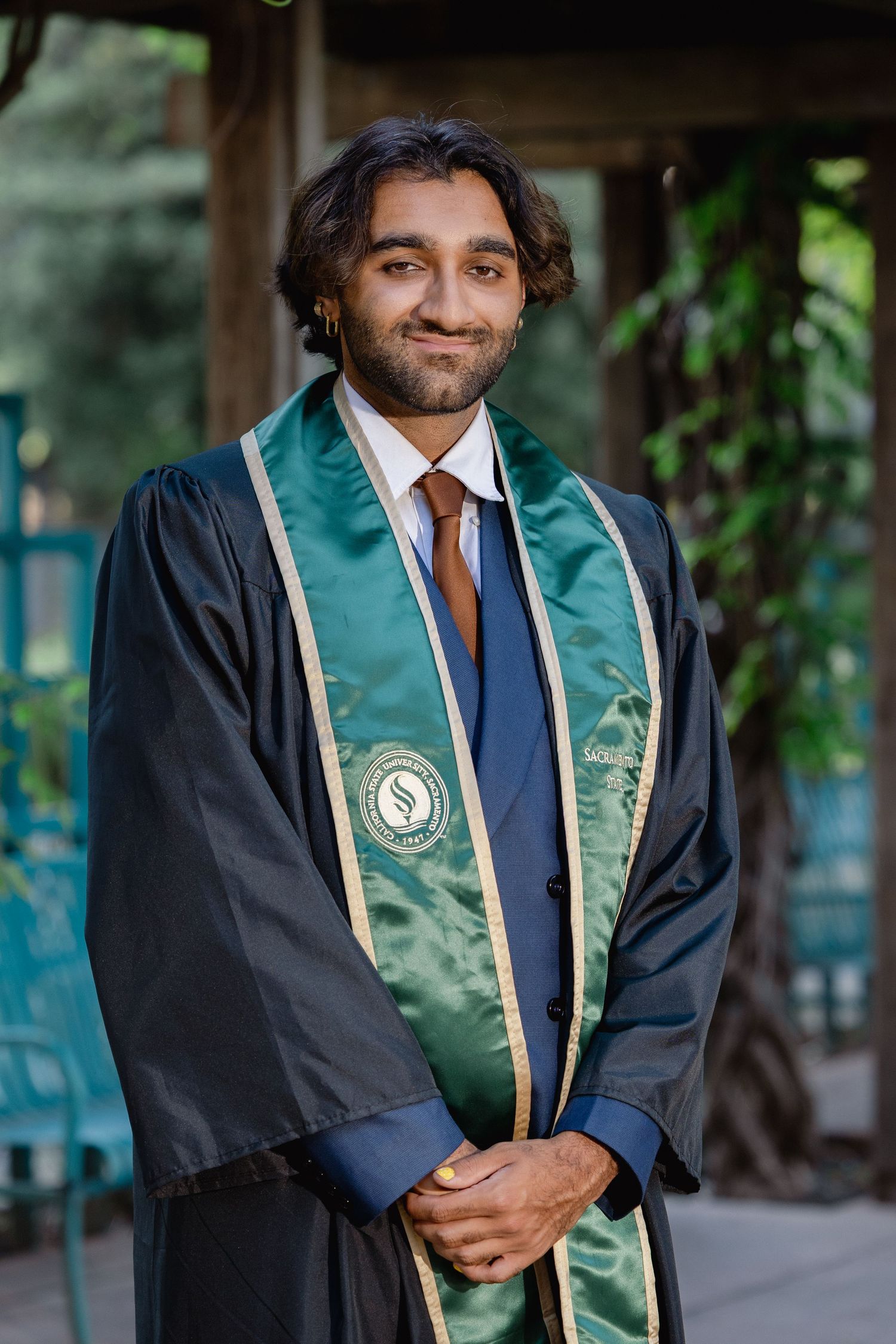 Young man in black graduation gown with green sash smiling outdoors.