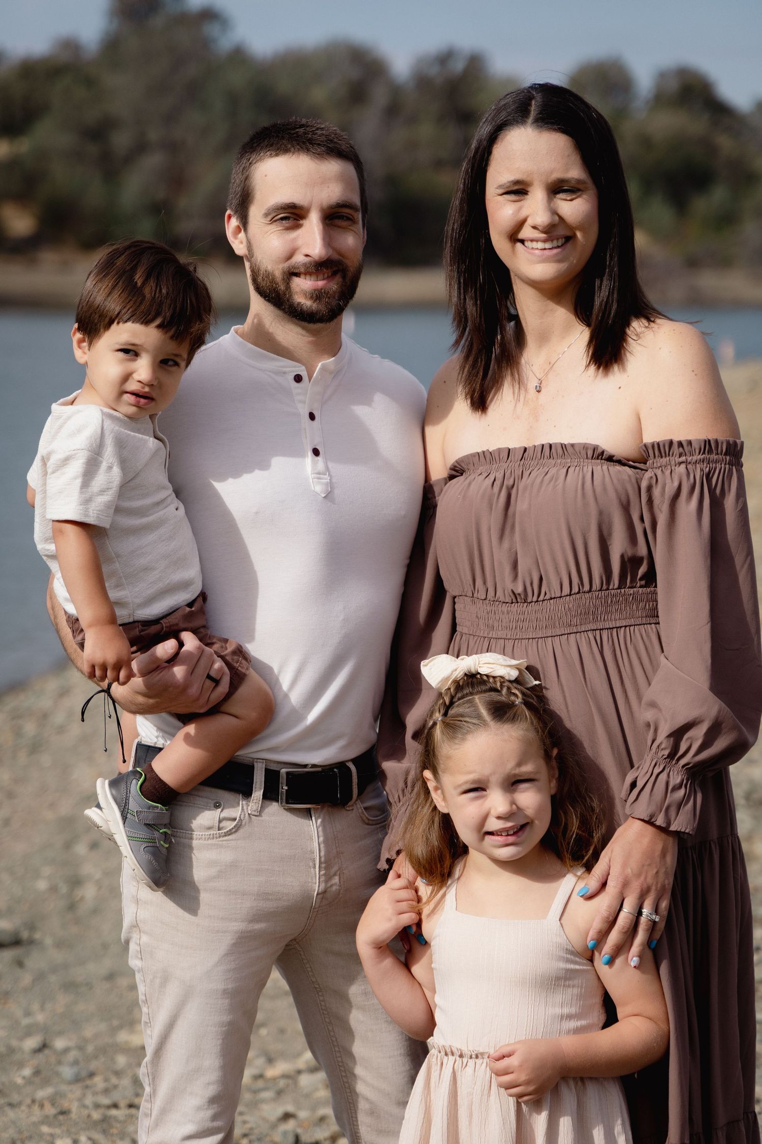 Family of four posing outdoors near a lake, parents smiling with two young children on a sunny day.