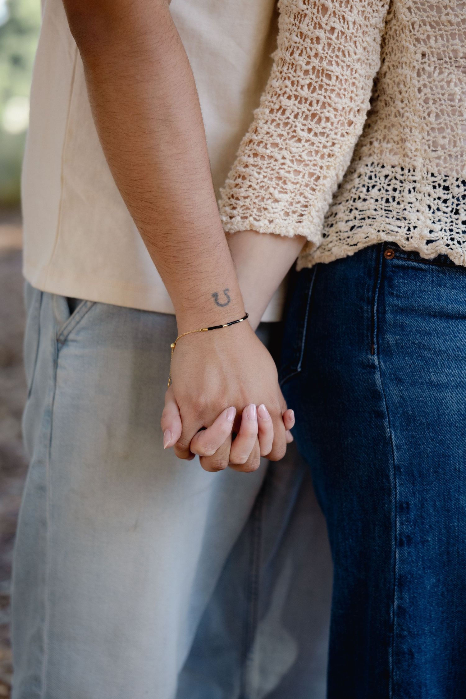Close-up of a couple holding hands, one with a horseshoe tattoo and bracelet, wearing casual outfits outdoors.
