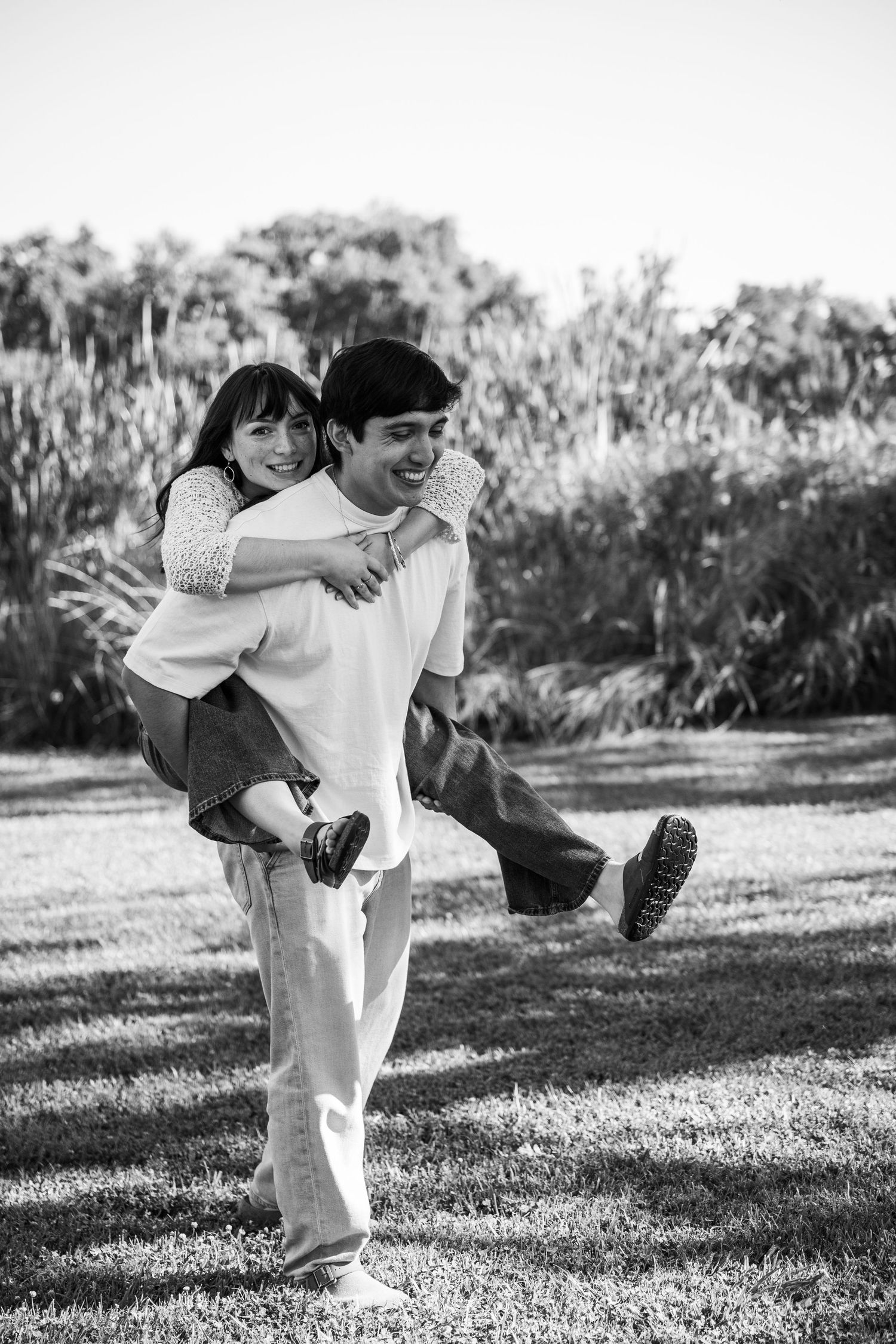 Black and white photo of a young man giving a smiling woman a piggyback ride outdoors on grass.