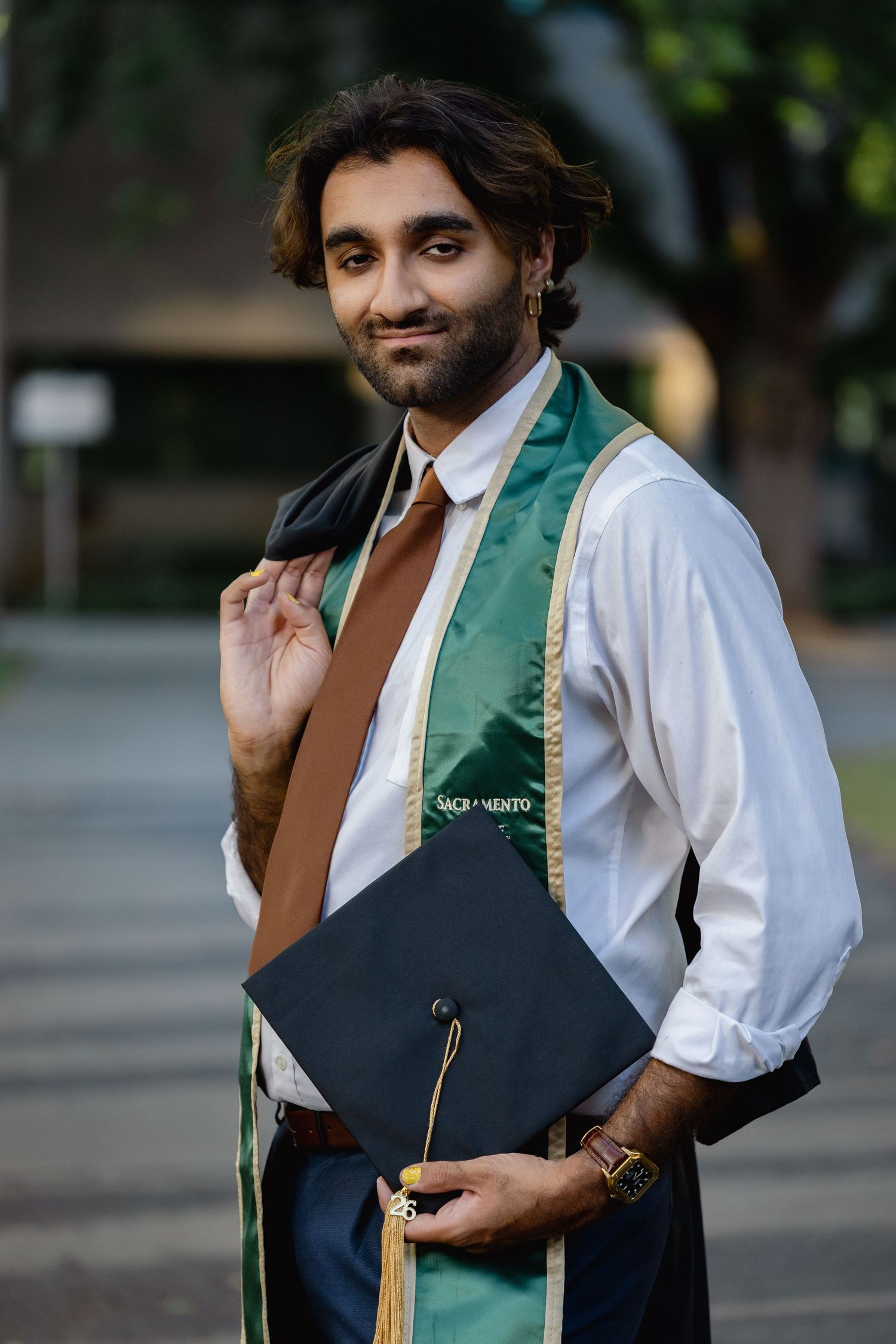 Smiling graduate holds cap while wearing green Sacramento State stole over white shirt and tie.