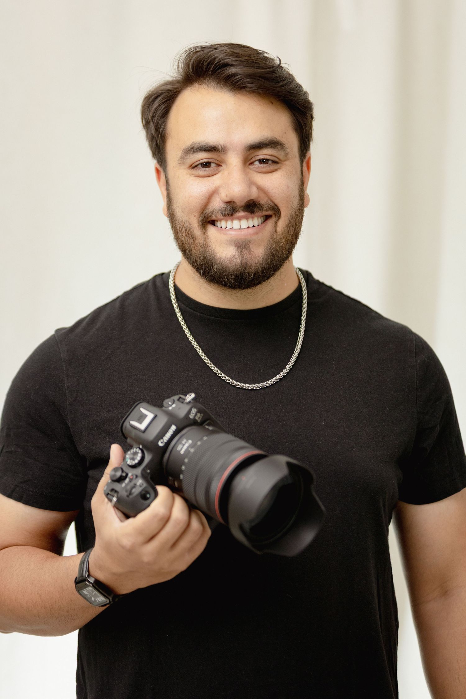 Smiling young male photographer holding a professional DSLR camera against a light background.
