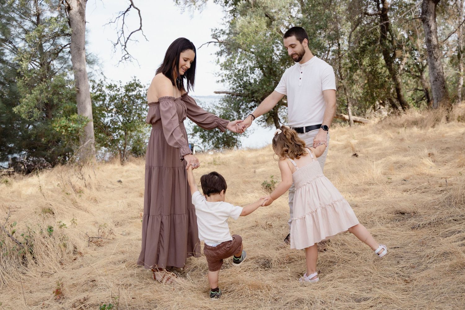 Family of four holding hands and playing in a golden field surrounded by trees during an outdoor photo session.