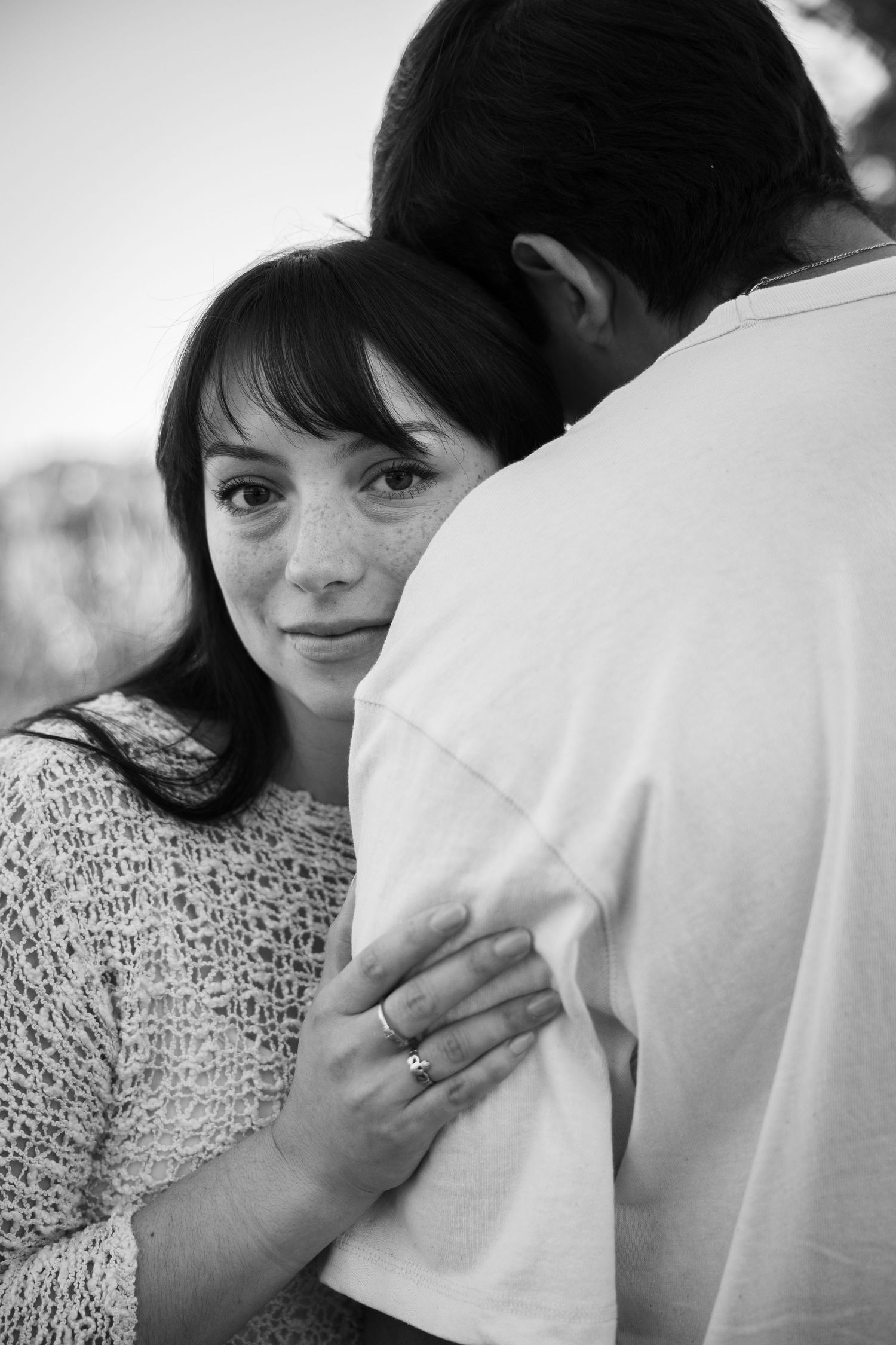 Black and white photo of a young couple embracing, woman with freckles resting her head on man's shoulder.