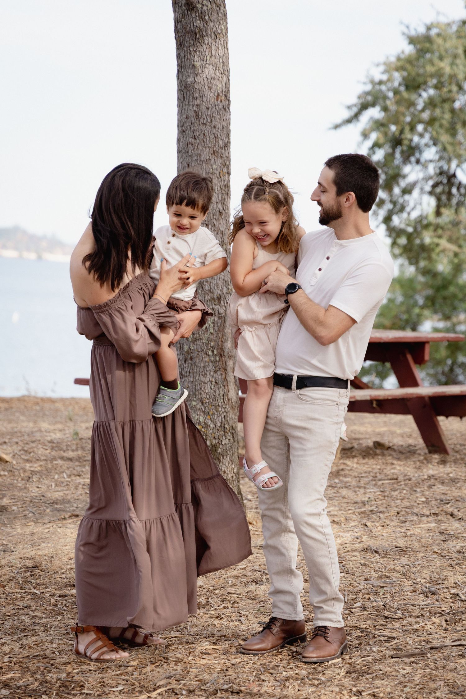 Family of four poses by a tree near a lake, parents holding young children in neutral-toned outfits on a sunny day.