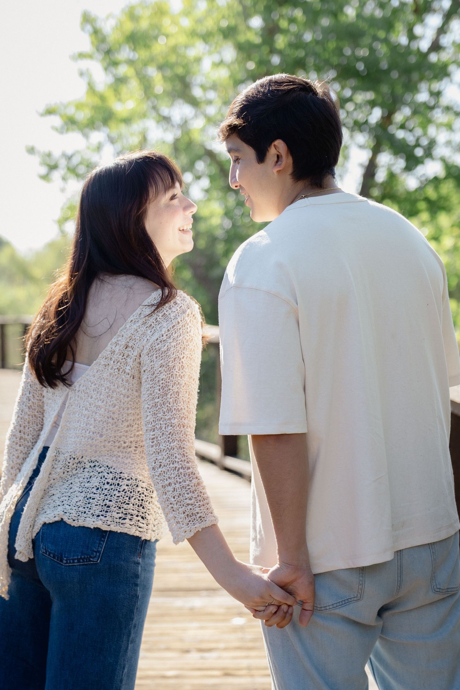 Young couple holding hands on a wooden bridge, smiling at each other surrounded by lush green trees.