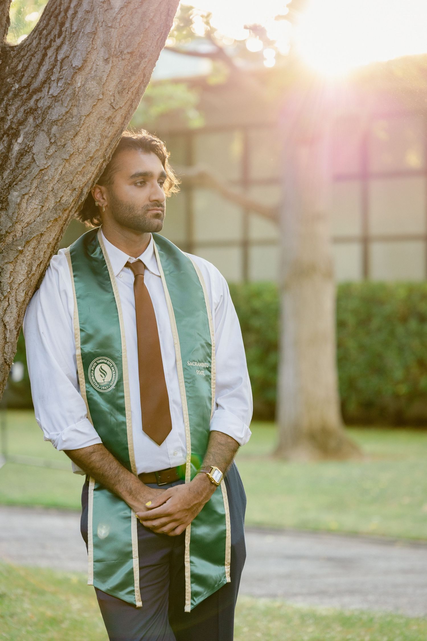 Young man in graduation stole and tie leaning against tree outdoors during golden hour.