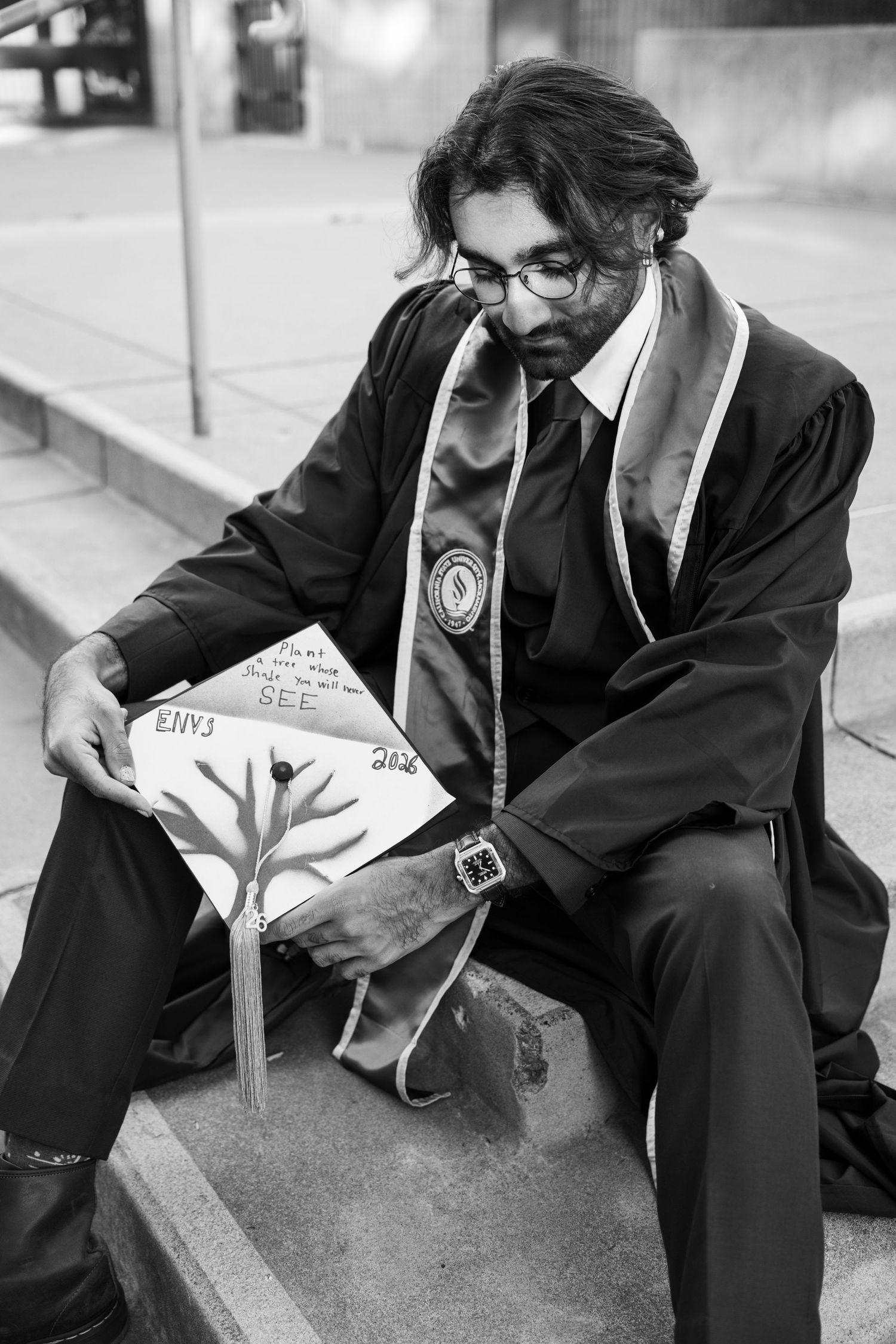 Graduate in black robes sits on steps holding decorated mortarboard cap in black and white photo.