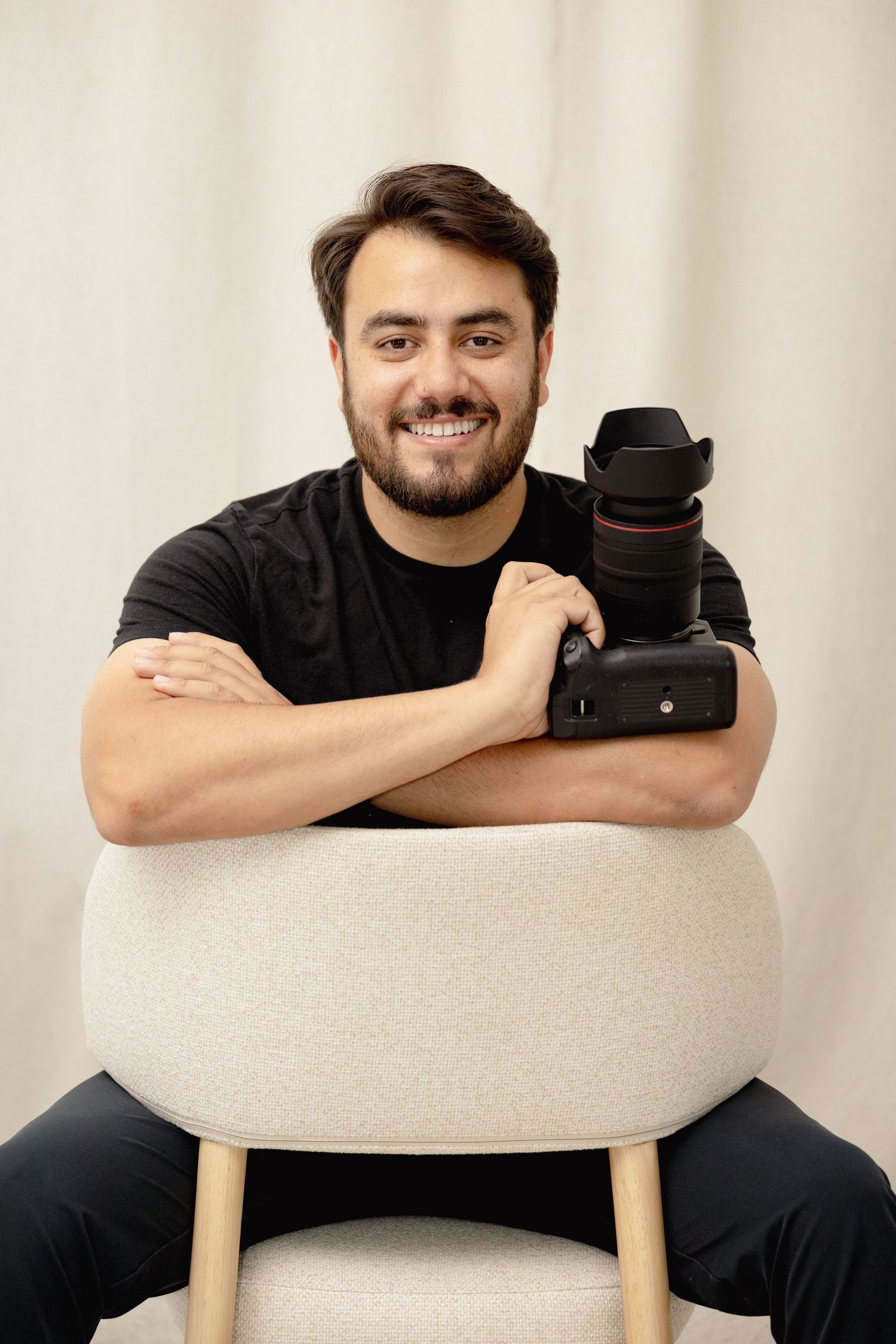 Smiling bearded photographer in black shirt posing with DSLR camera on chair indoors.