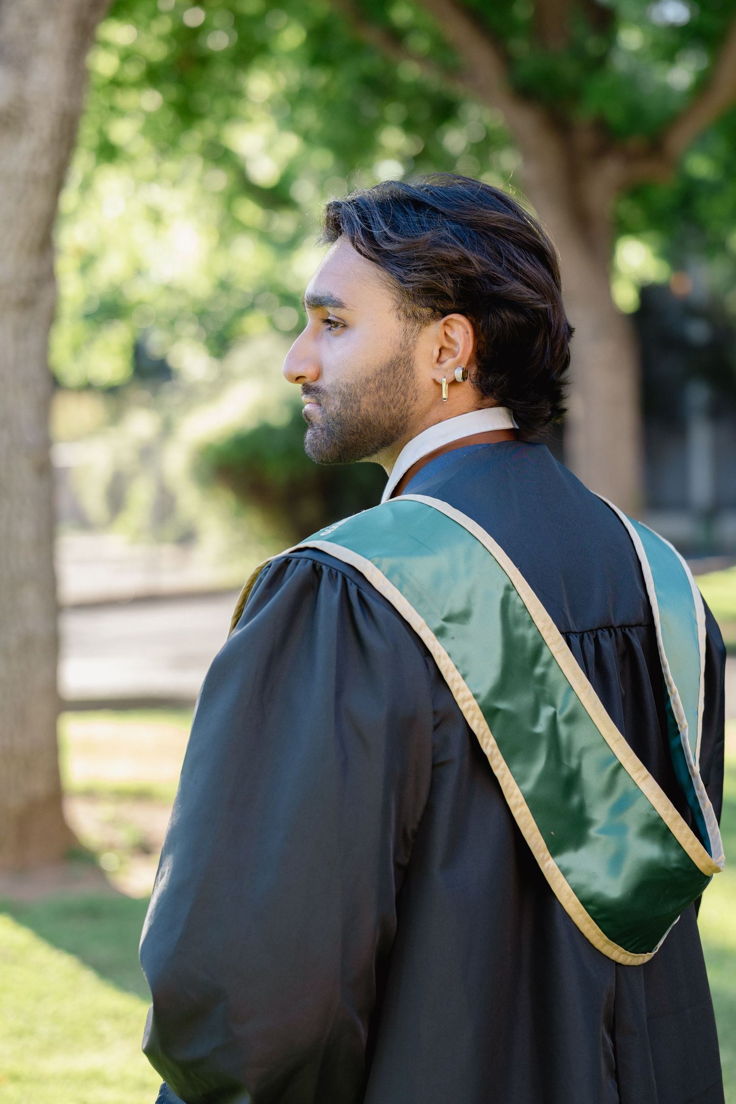 Young man in black and green graduation gown standing outdoors near trees on a sunny day.