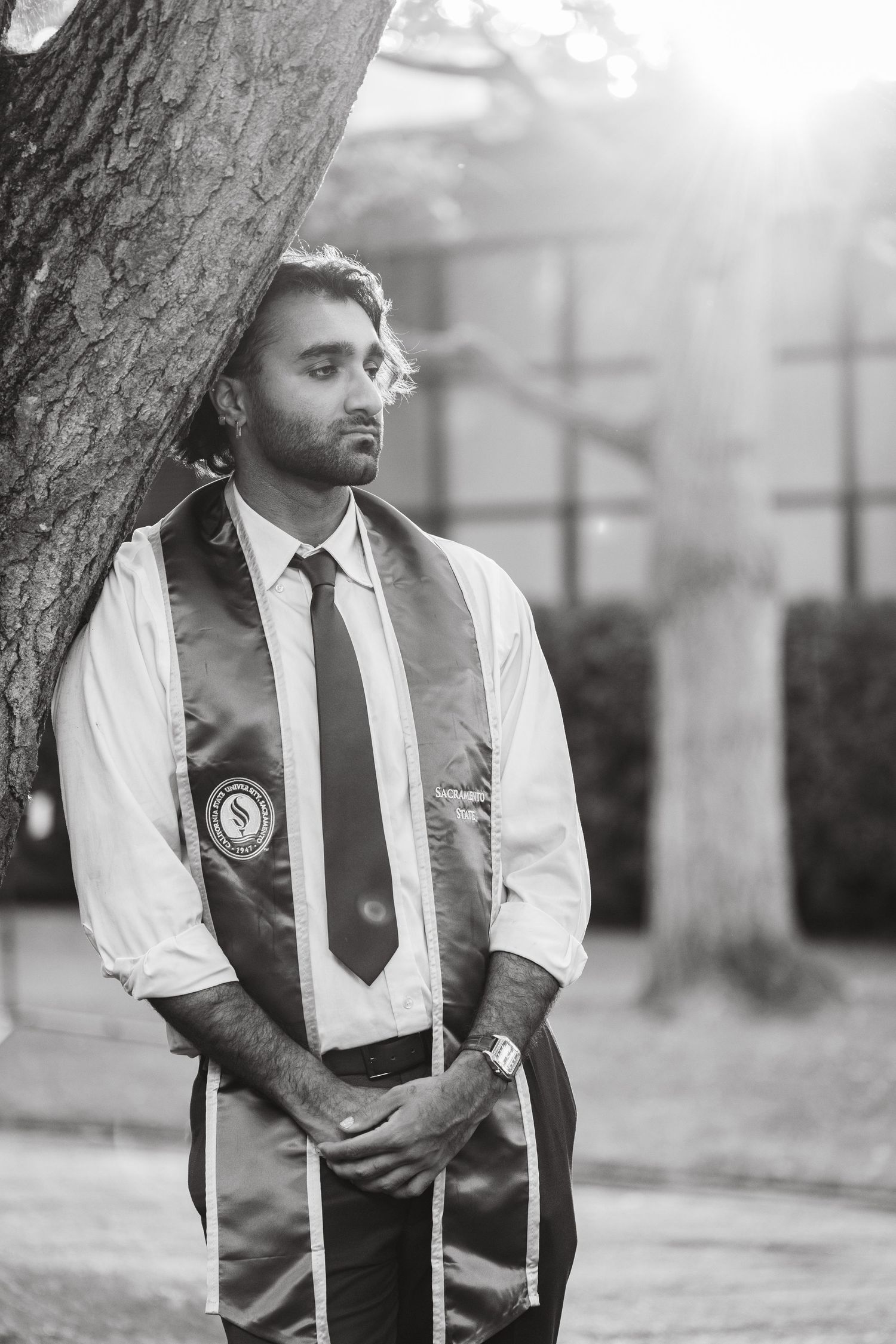Graduate in white shirt and stole leans against tree in black and white photo.