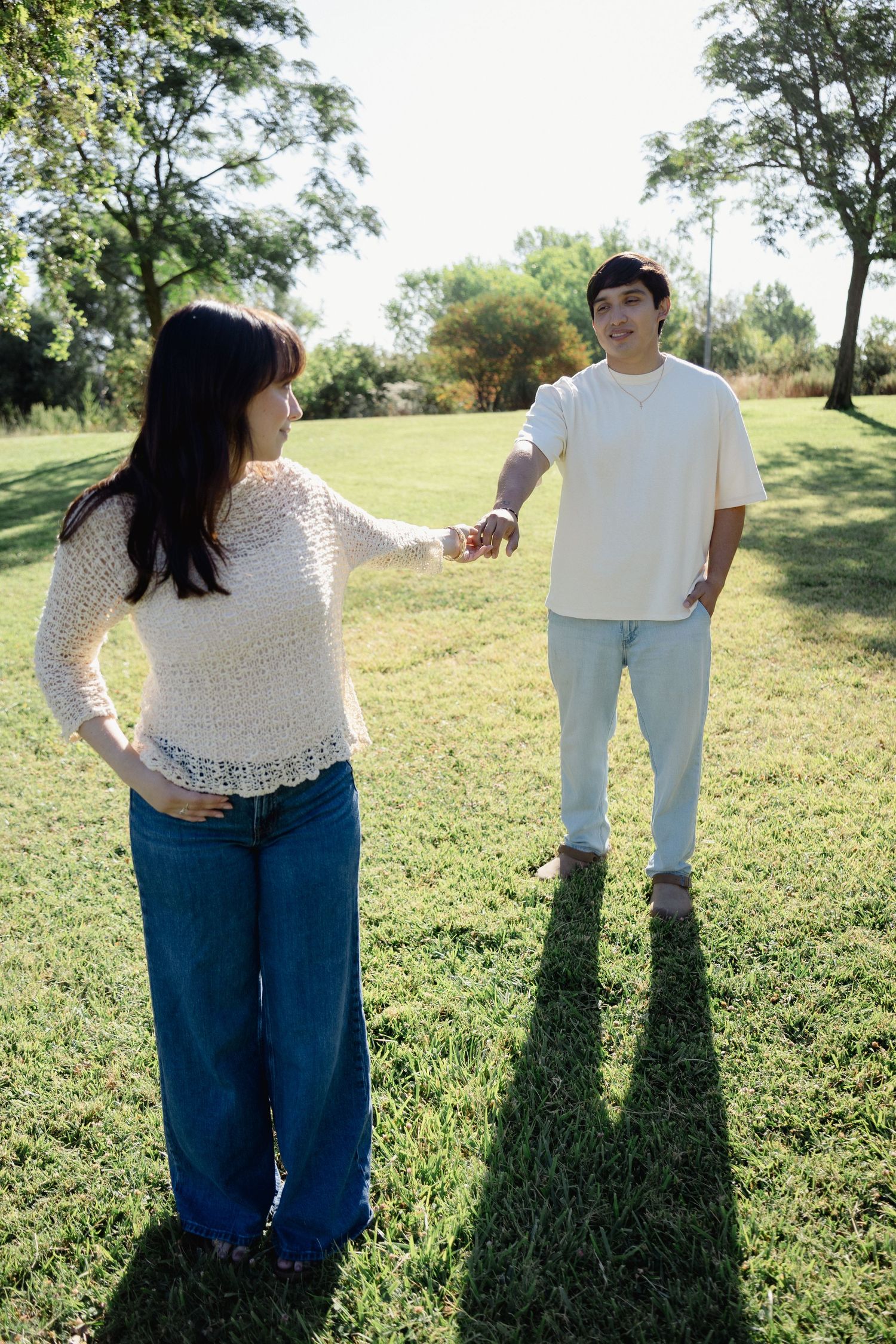 Young couple holding hands in a sunny park, standing on green grass with trees in the background.