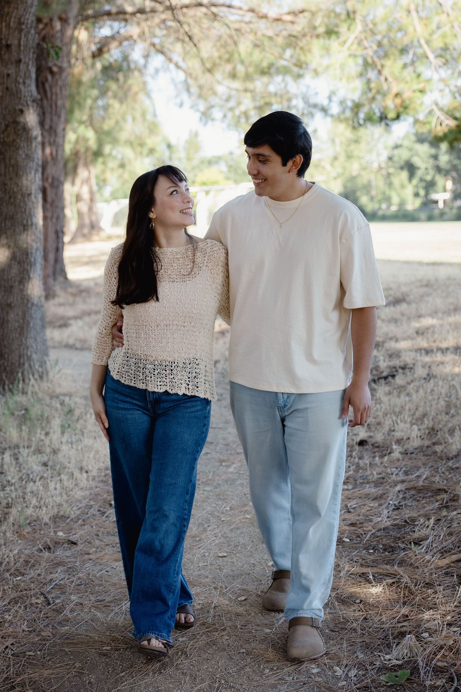 Young couple walking together on a shaded park path, smiling at each other in casual summer outfits.