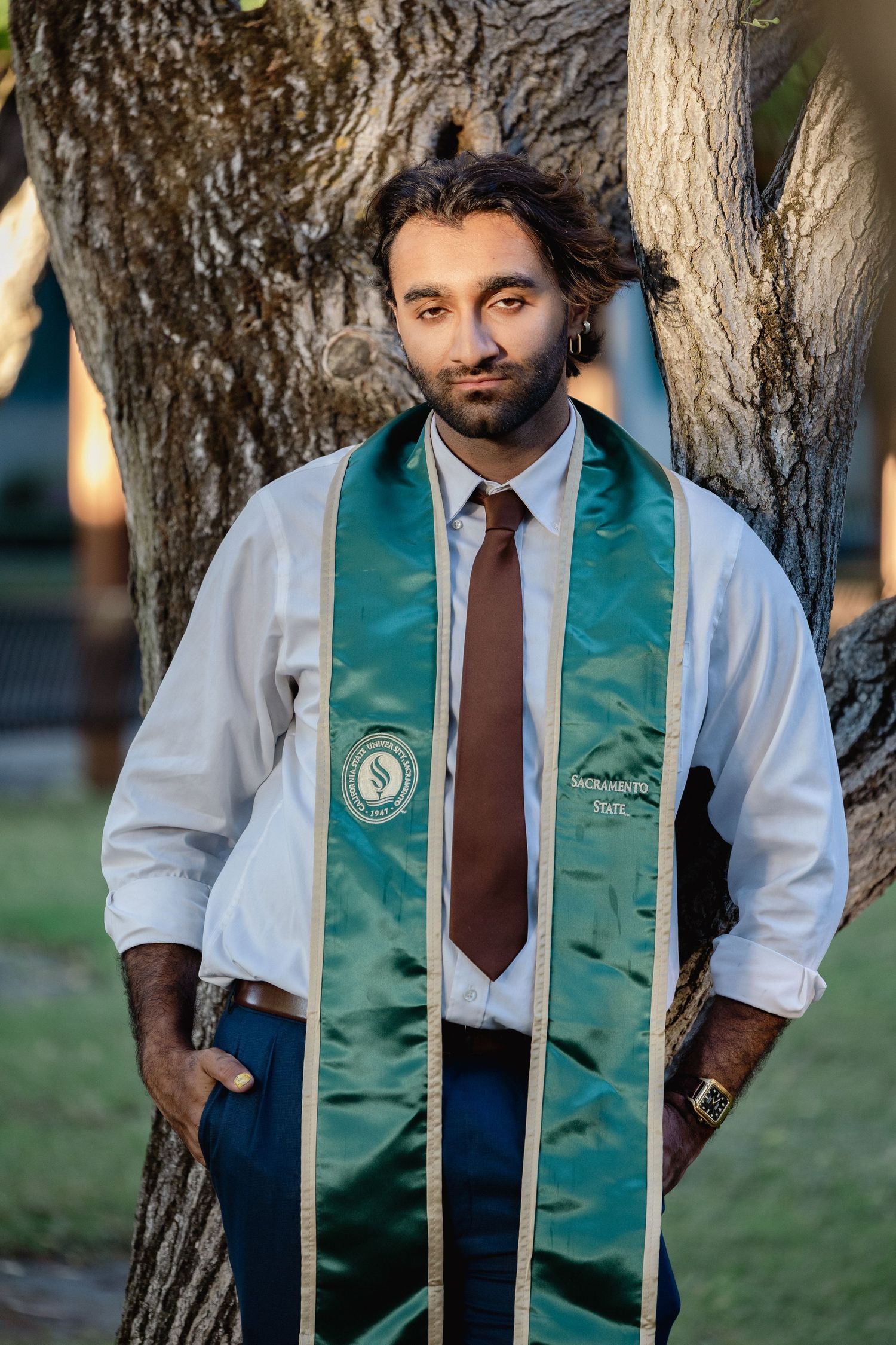 Graduate in white shirt and green Sacramento State stole leans against tree outdoors.